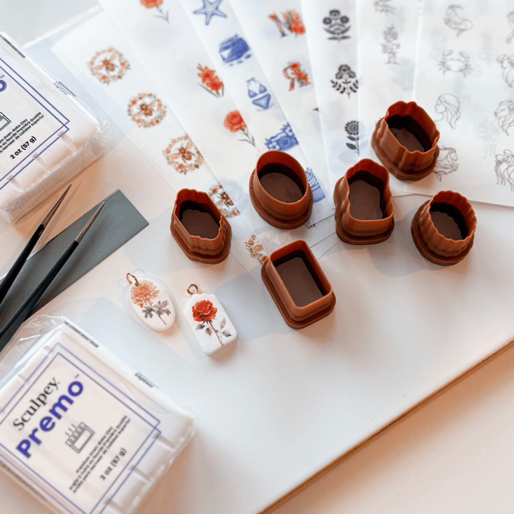 Claying supplies including small molds, decorative charms, paintbrushes, stamps, and patterned paper on a white workspace.