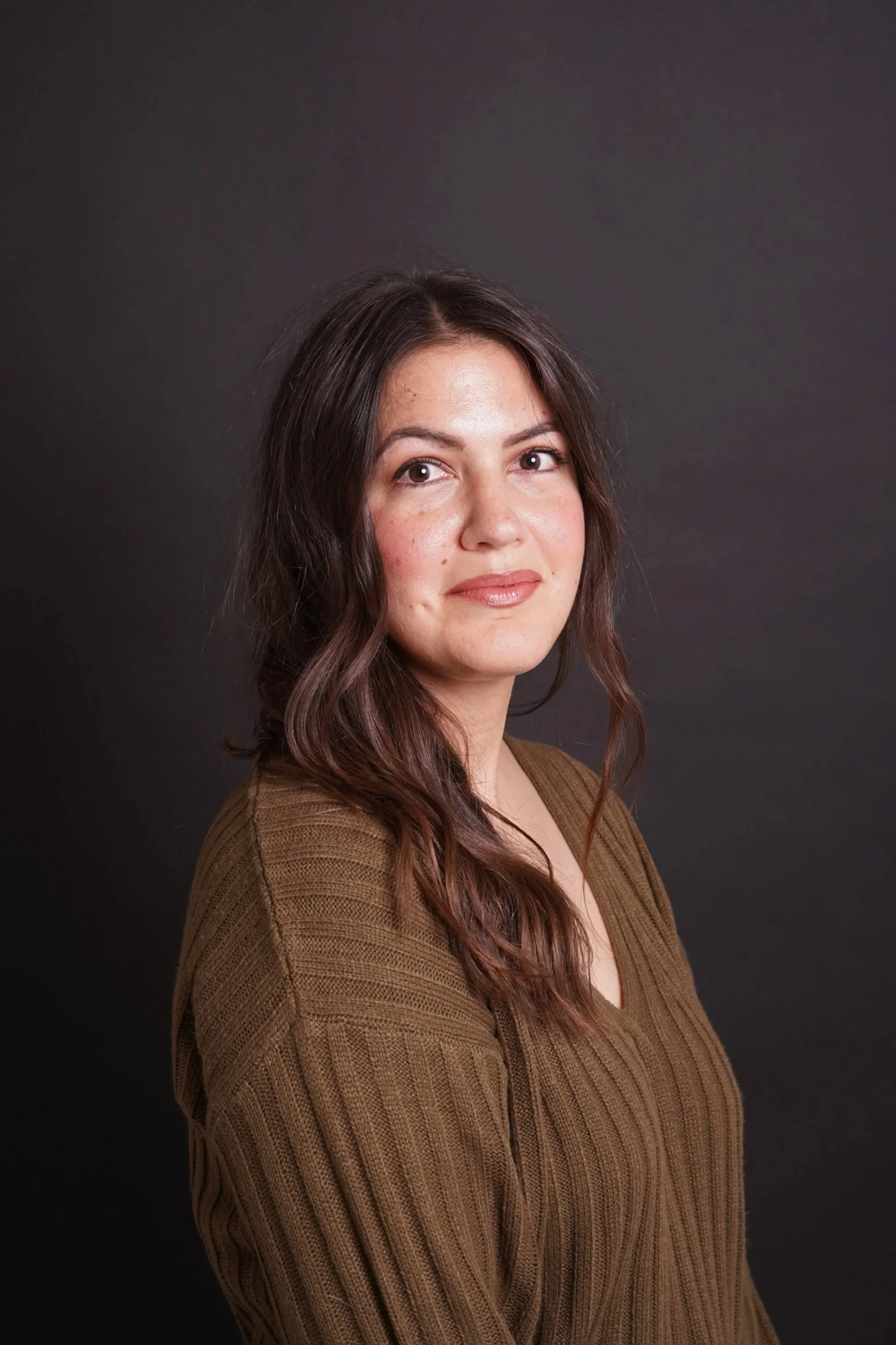 A woman with long, wavy brown hair and light skin, wearing a brown ribbed sweater, standing against a dark background.