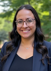 Portrait of a woman with long dark hair, wearing glasses, a navy blazer, and a necklace, smiling outdoors with green trees in the background.