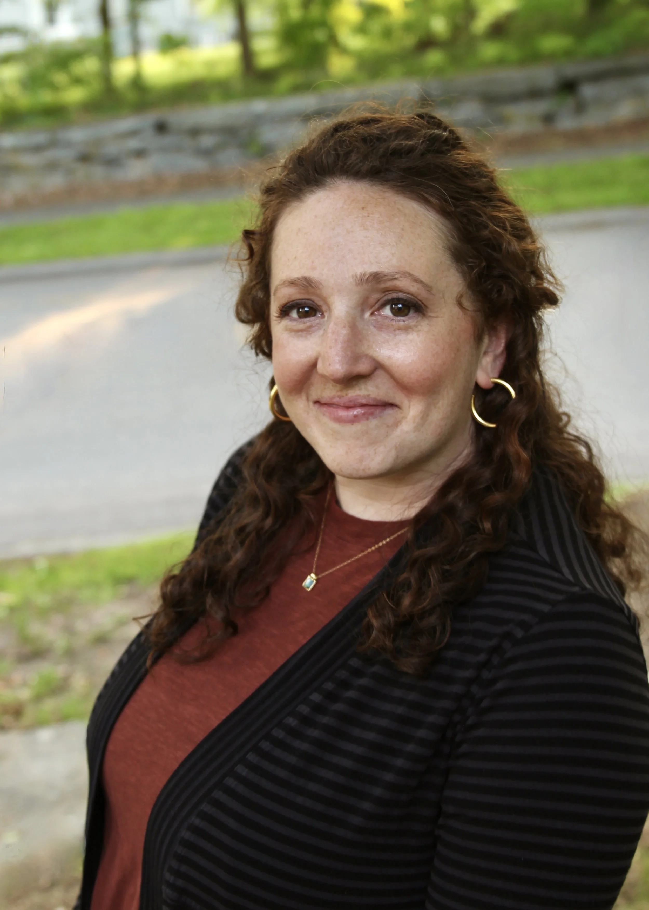 A woman with curly brown hair smiling outdoors, wearing gold hoop earrings, a gold necklace with a small pendant, and a black jacket over a dark red shirt, with trees and a grassy area in the background.