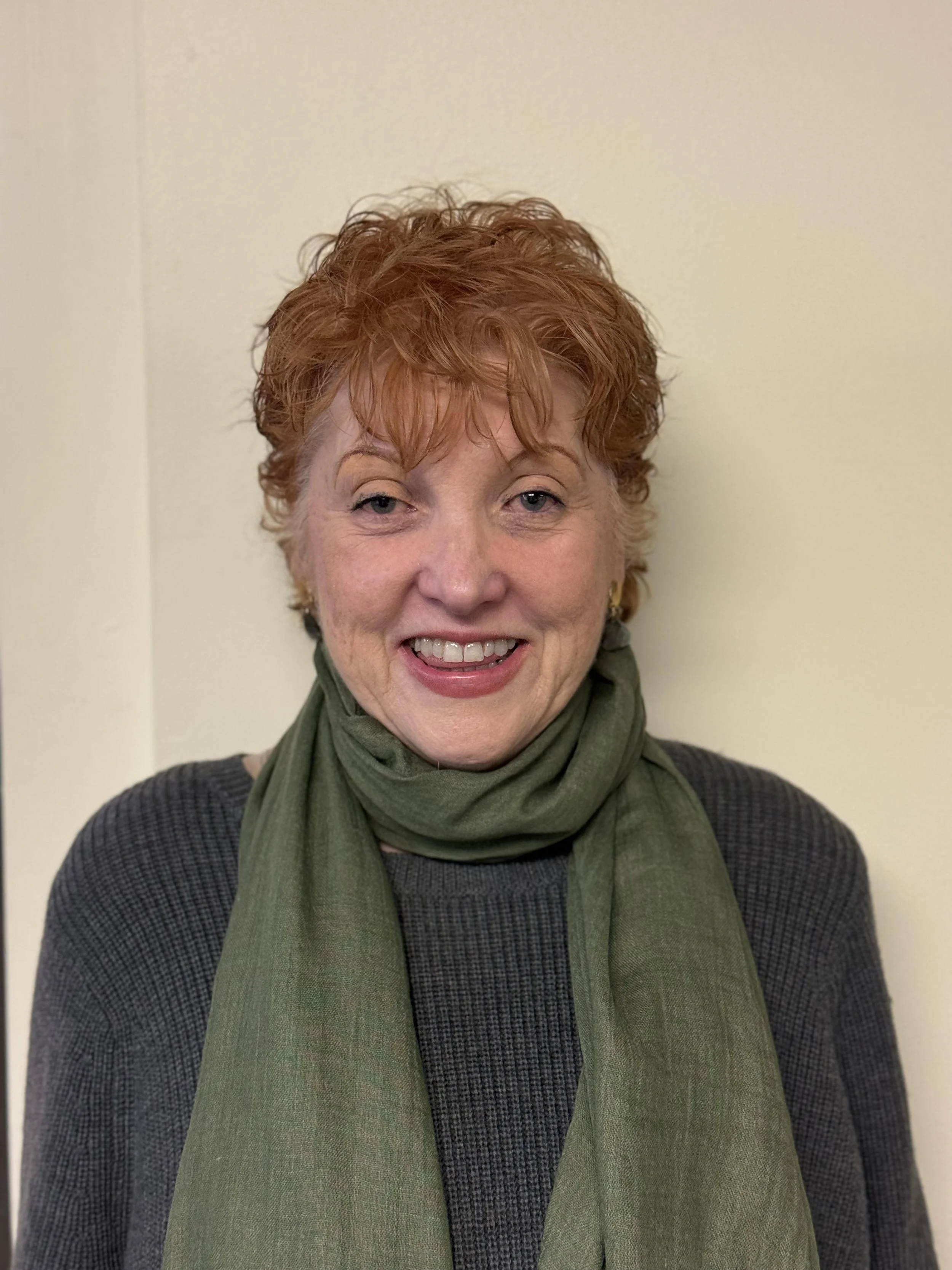 A woman with short, curly red hair smiling, wearing a dark gray sweater and an olive green scarf against a plain light-colored wall.