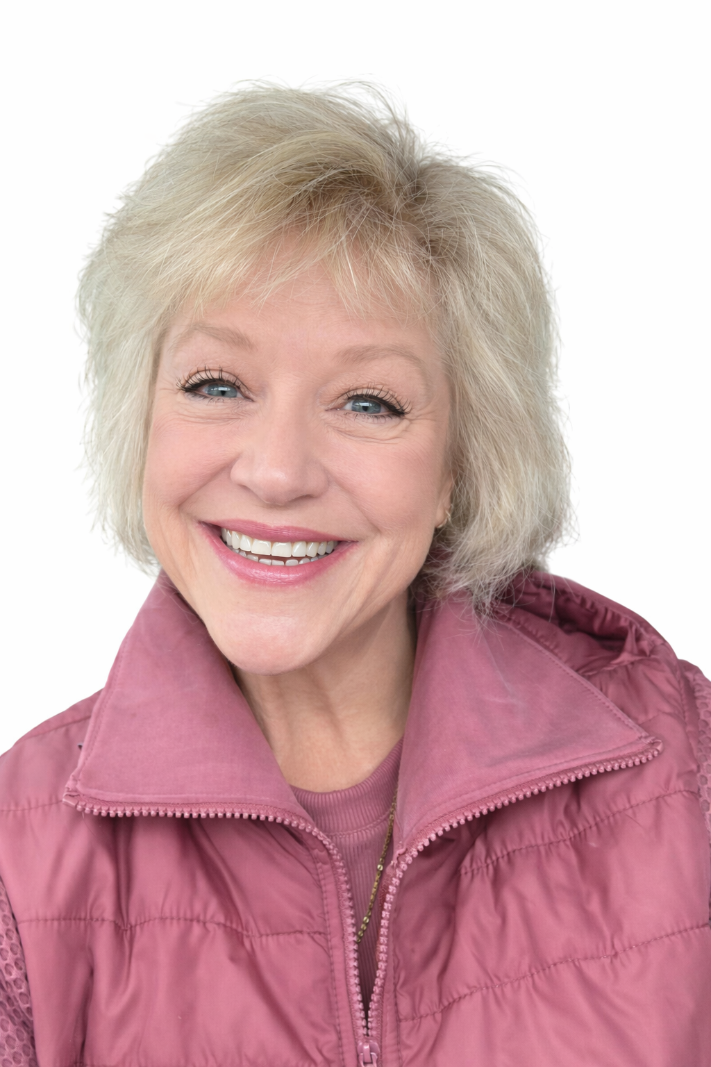 A smiling elderly woman with short, blonde hair and bright blue eyes, wearing a pink jacket and a pink shirt, against a plain white background.