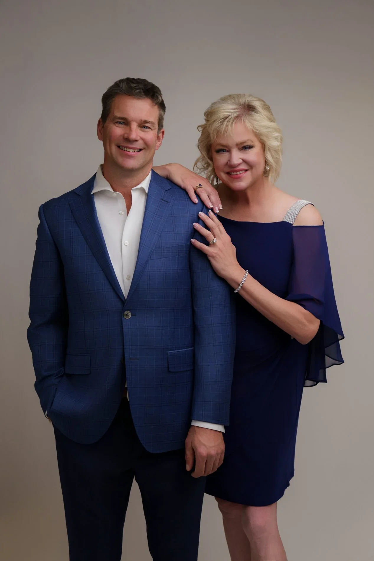 A man and a woman standing close together, smiling at the camera, against a plain background. The man is wearing a blue suit jacket and white shirt, and the woman is wearing a navy blue dress with sheer sleeves.