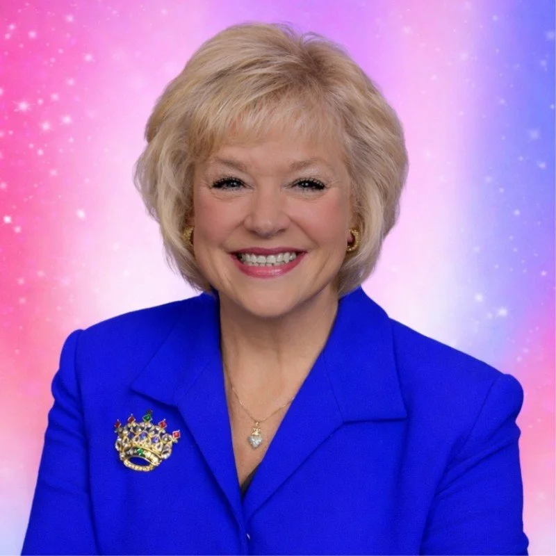 A smiling woman with blonde hair wearing a royal blue blazer and a crown-shaped brooch, standing against a pink and blue starry background.