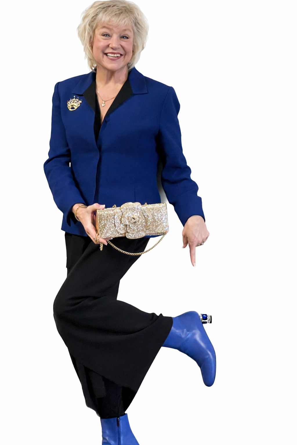 A woman with blonde hair smiling, wearing a blue blazer with a crown pin, black pants, and blue boots, holding a decorative gold clutch purse with a chain strap, standing against a white background.