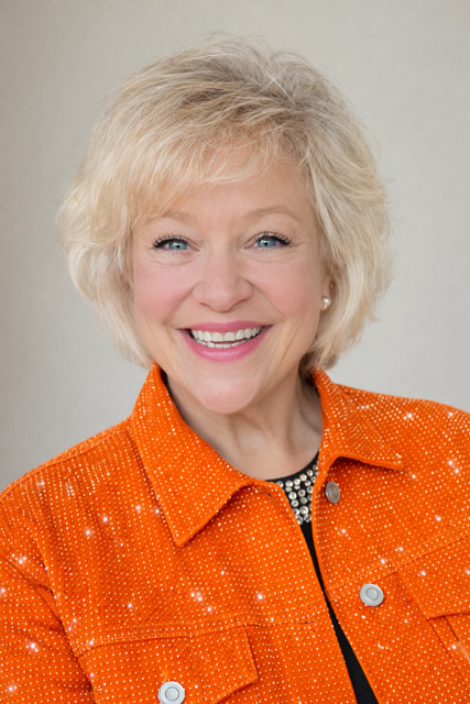 Smiling woman with blonde hair wearing an orange jacket with silver buttons and a black top with white patterns in front of a light neutral background.