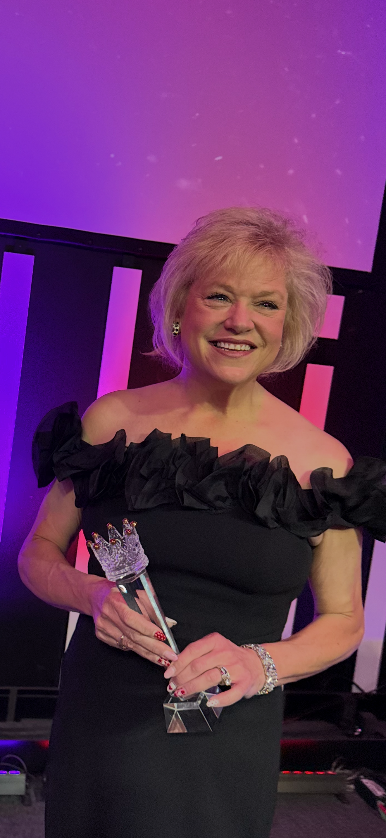 A woman with blonde hair in a black dress with ruffled shoulders holding a crystal trophy with a crown on top, smiling at an event with colorful lighting.