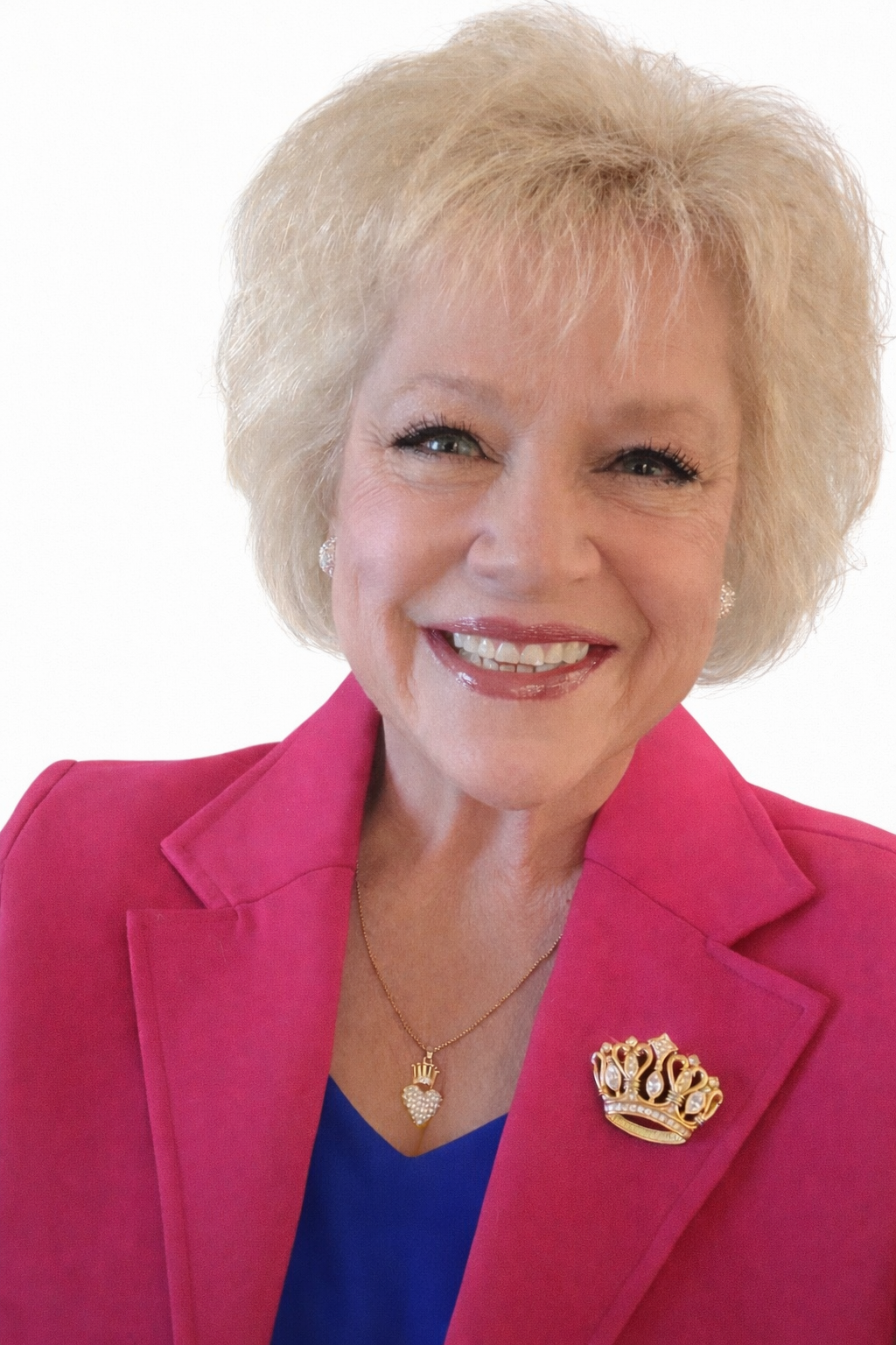 Smiling middle-aged woman with short blonde hair, wearing a pink blazer with a crown-shaped brooch, a necklace with a heart-shaped pendant, and pearl earrings.