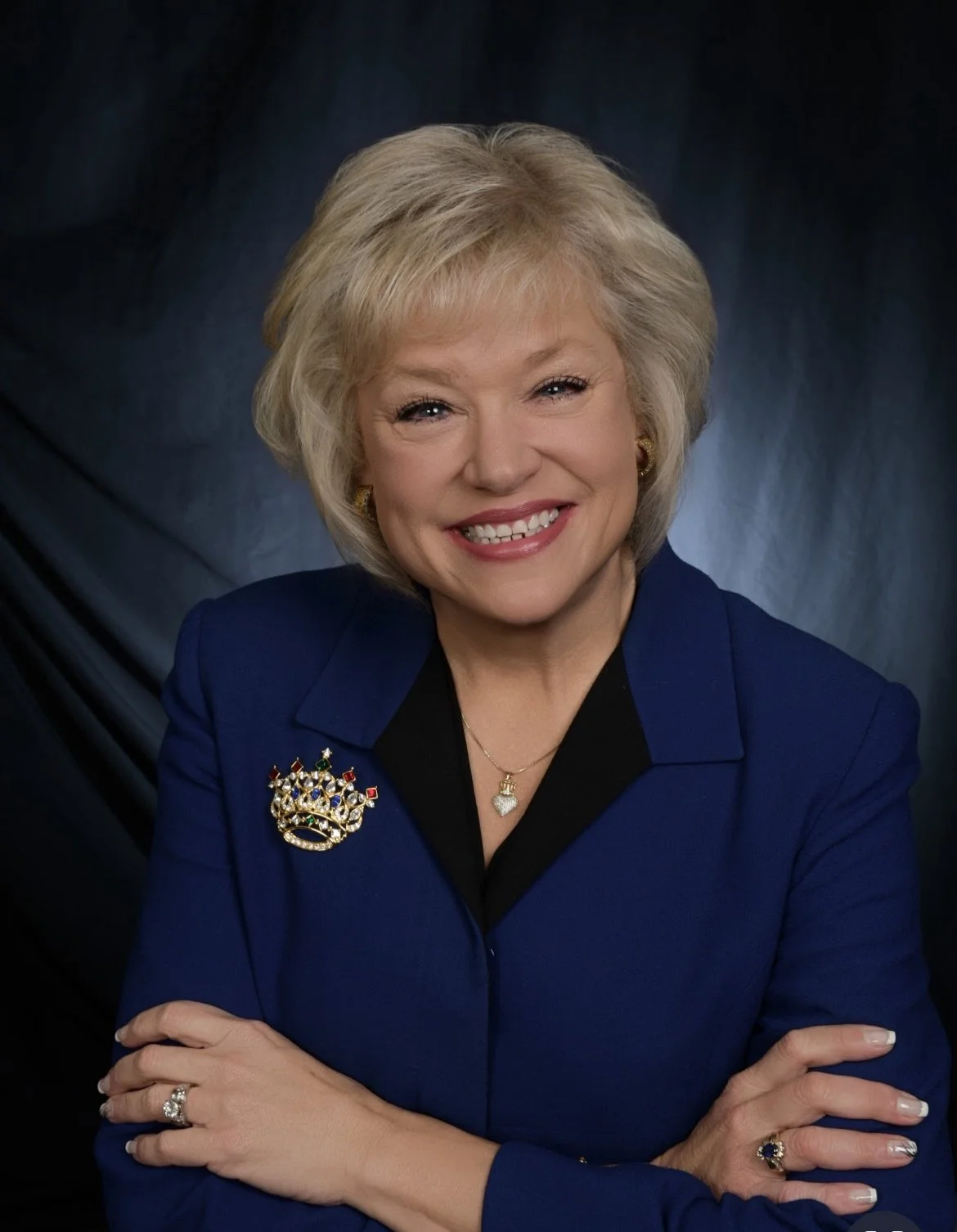 A woman with blonde hair smiling, wearing a royal blue blazer with a brooch, gold earrings, and a necklace, standing against a dark background.