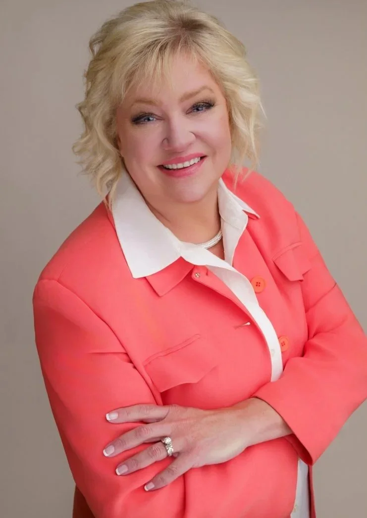 A middle-aged woman with blonde, curly hair smiling at the camera. She is wearing a coral-colored jacket over a white collared shirt and a pearl necklace. She has her arms crossed and is standing against a plain light gray background.