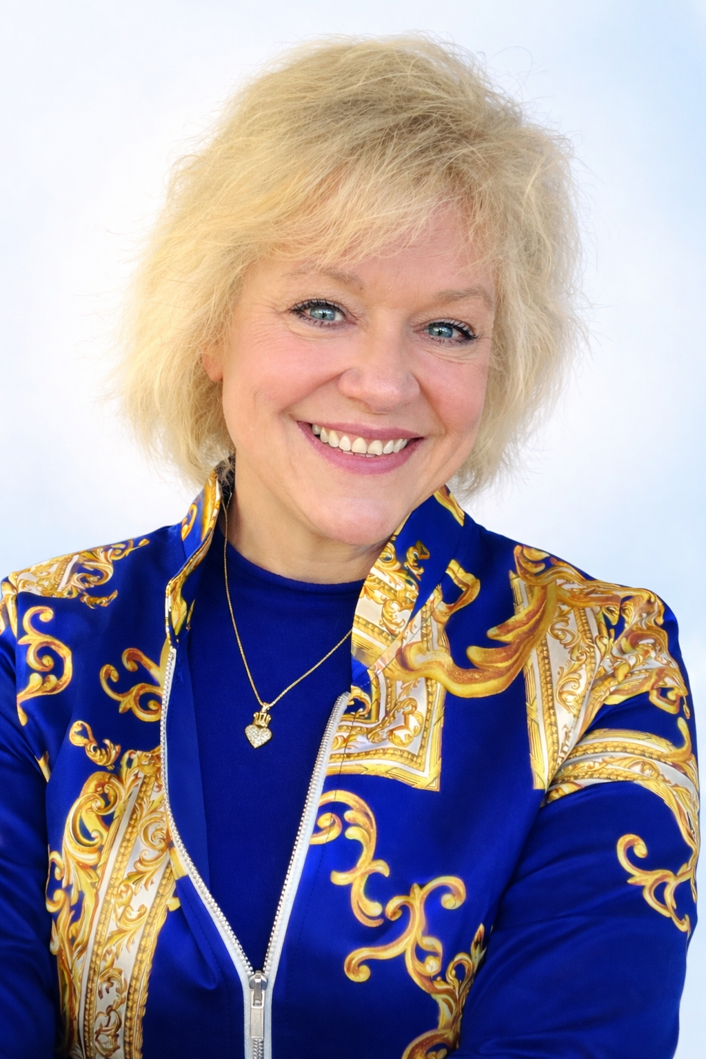 Smiling woman with blonde curly hair, wearing a blue and gold patterned jacket and a gold necklace with a heart-shaped pendant.