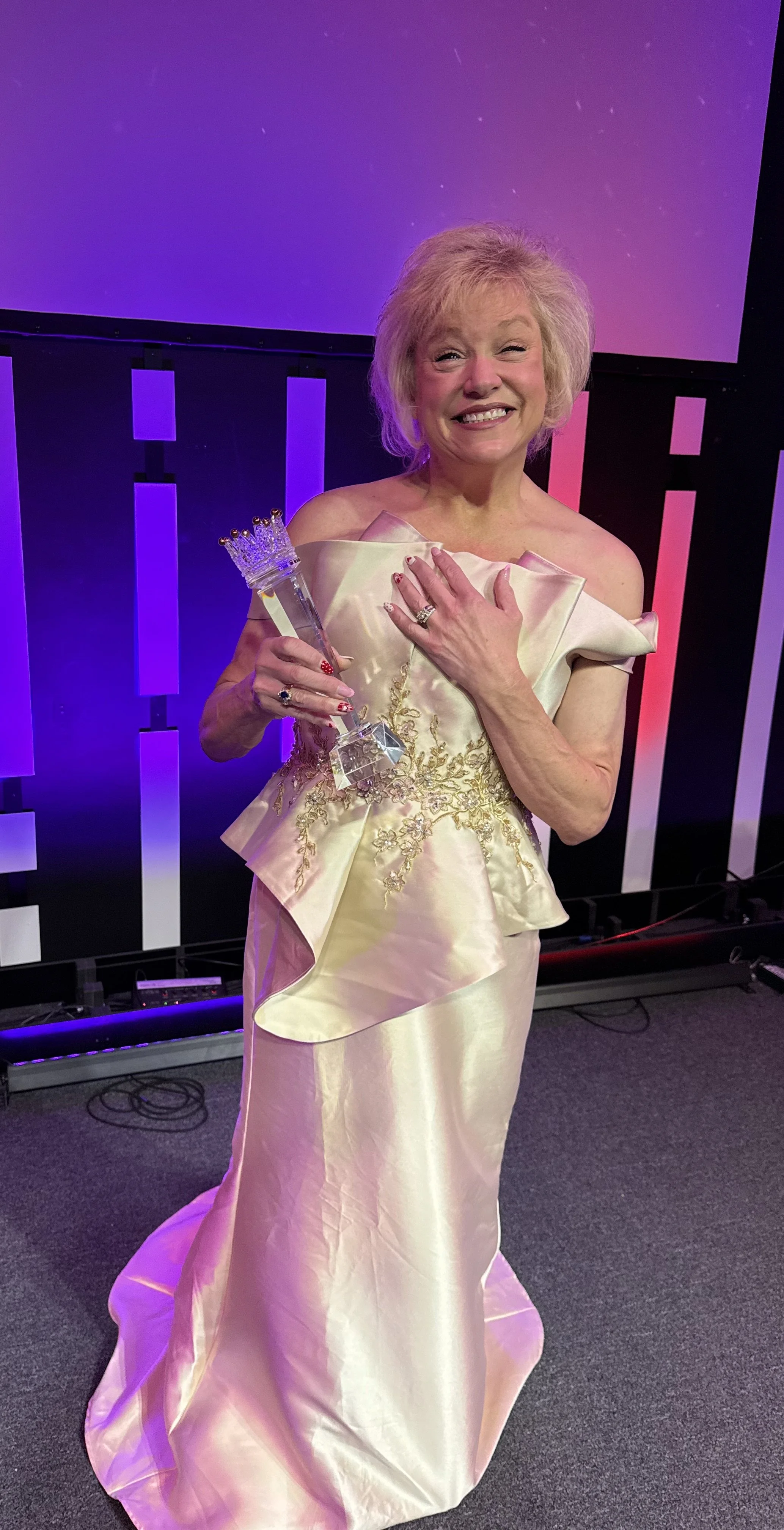 An older woman in a cream-colored satin gown holding a crystal trophy and smiling at an award ceremony.