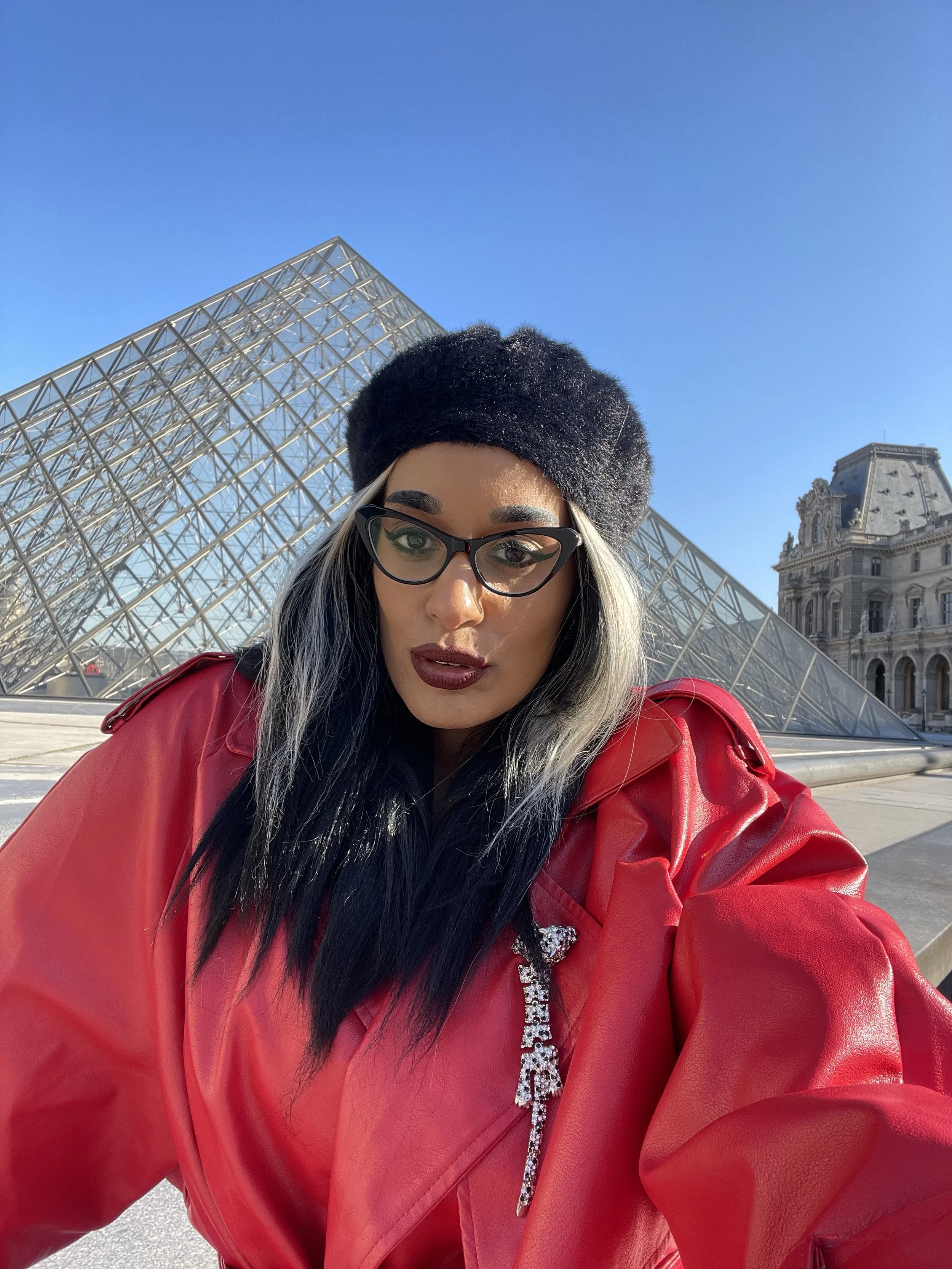 Roxy Murray taking a selfie at the Louvre Museum in Paris, wearing a black hat, glasses and red jacket, with the iconic glass Pyramid visible behind her