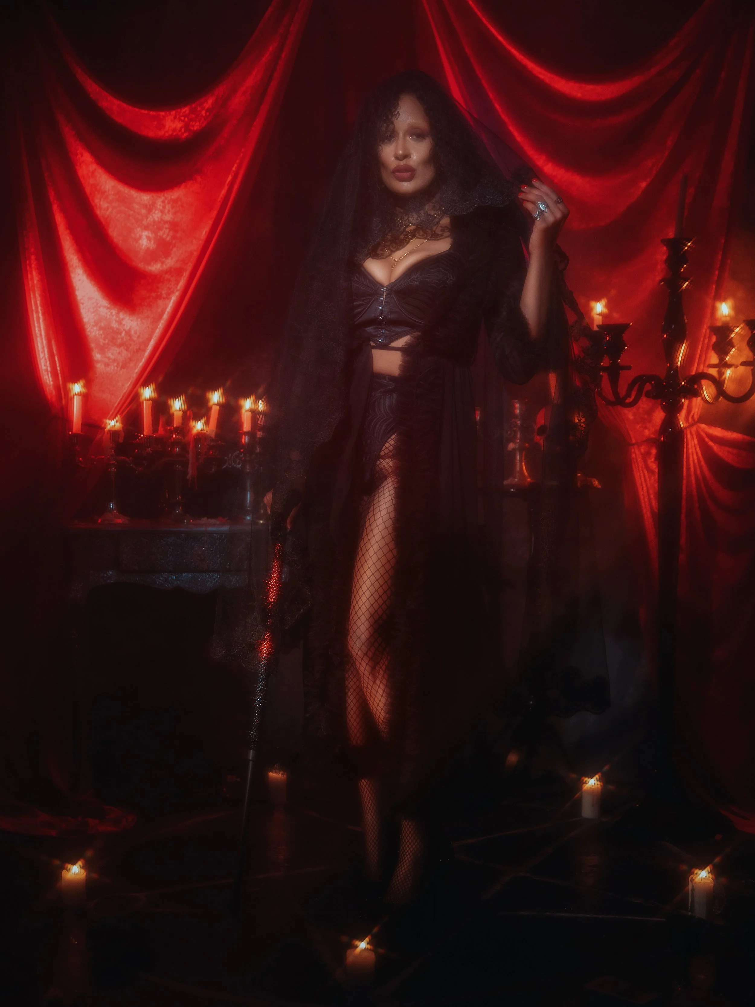 A woman dressed in black, standing in front of red velvet curtains with candles around her. She has dark curly hair, lace clothing, fishnet stockings, and a ring, giving a gothic appearance.
