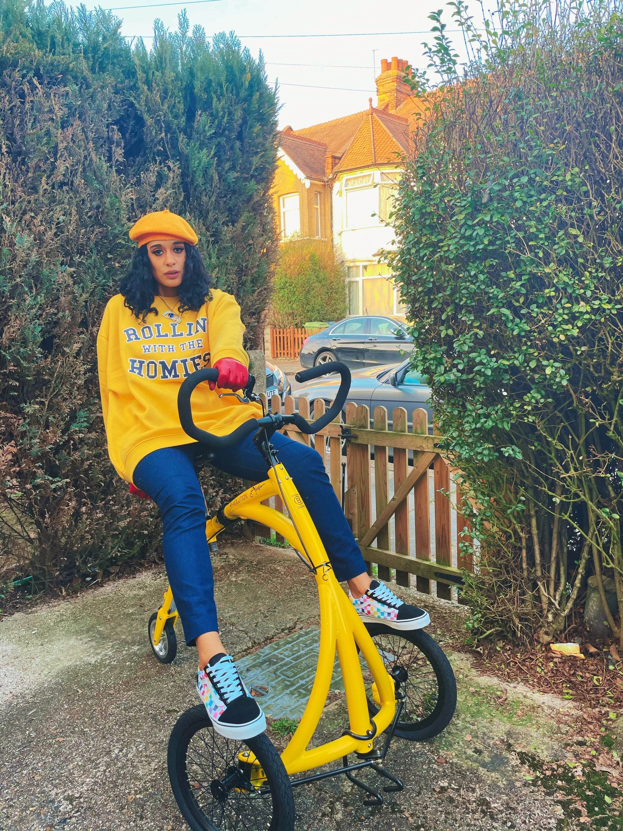 A young woman with dark, curly hair, wearing a yellow sweatshirt with copyrighted text, a yellow beret, red gloves, and colorful sneakers, sits on a yellow electric bike outdoors near a wooden fence and behind dense greenery, with houses and parked cars visible in the background.