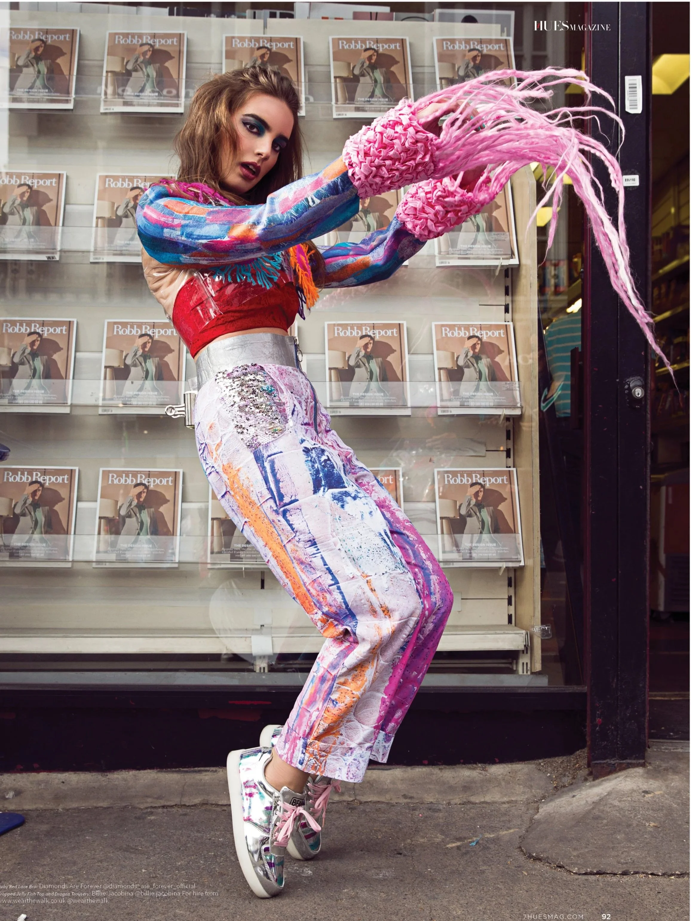 A young woman with long, wavy brown hair wearing multicolored, tie-dye style pants, a rainbow-colored sweater, and shiny sneakers is standing in front of a magazine display, posing with her arm extended towards the camera.
