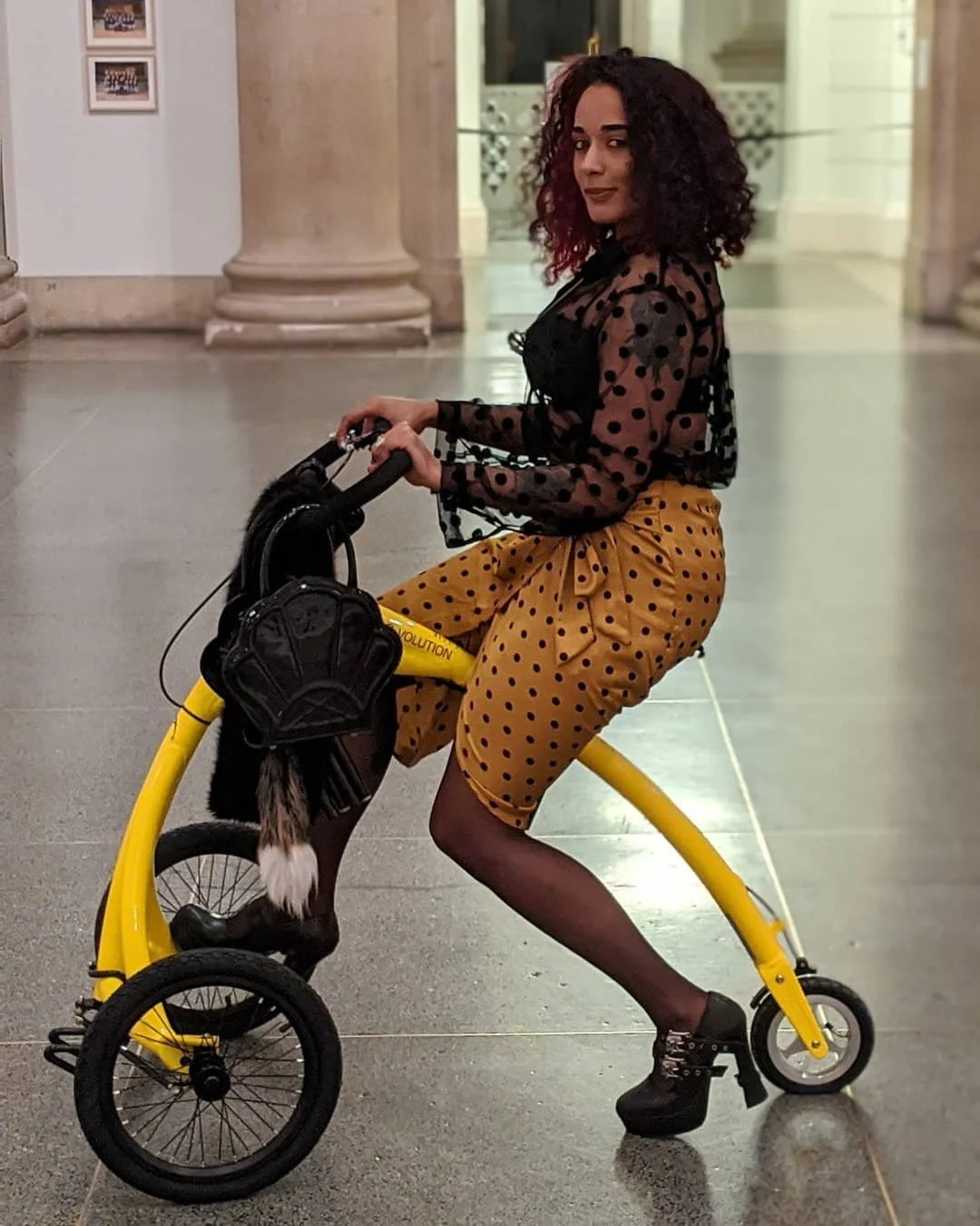 A woman with curly hair wearing a black sheer polka dot blouse, yellow polka dot pants, and high heel shoes, sitting on a yellow miniature bicycle in an indoor setting with columns and framed pictures on the wall.