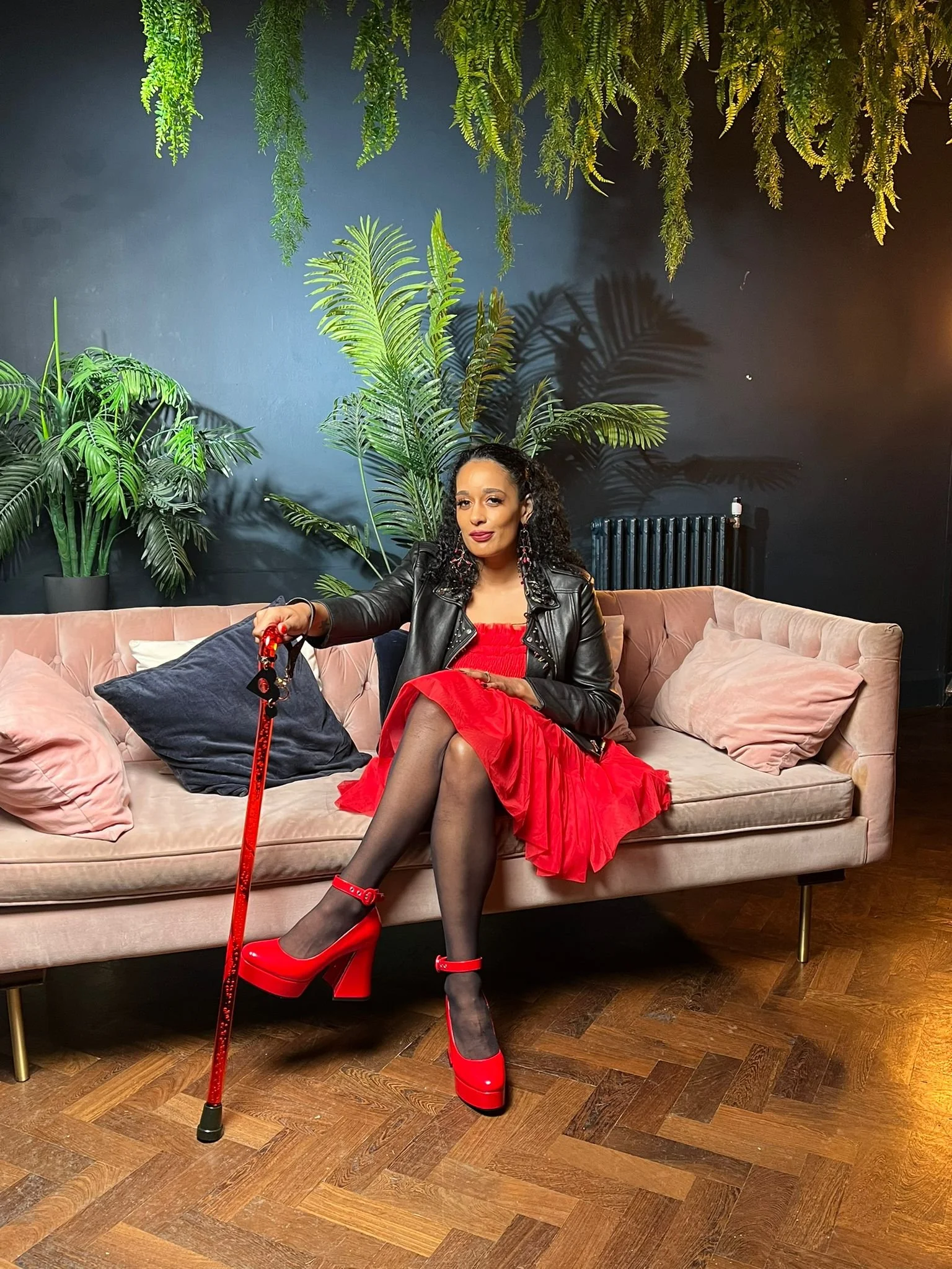 A woman sitting on a light pink sofa in a room with dark walls and large green plants. She is wearing a red dress, black stockings, red high heels, and a black leather jacket. She holds a red leash attached to a prosthetic leg and has curly hair and earrings.