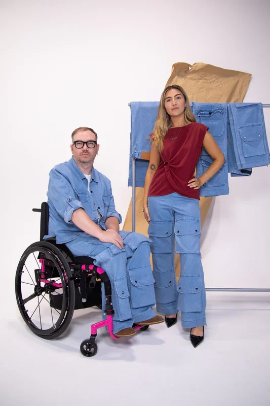A man in a wheelchair and a woman standing next to him pose in front of a plain white background. The man wears glasses and a light blue jumpsuit, and the woman is dressed in a red top and blue cargo pants. Behind them is a clothing rack with blue and brown fabric items.