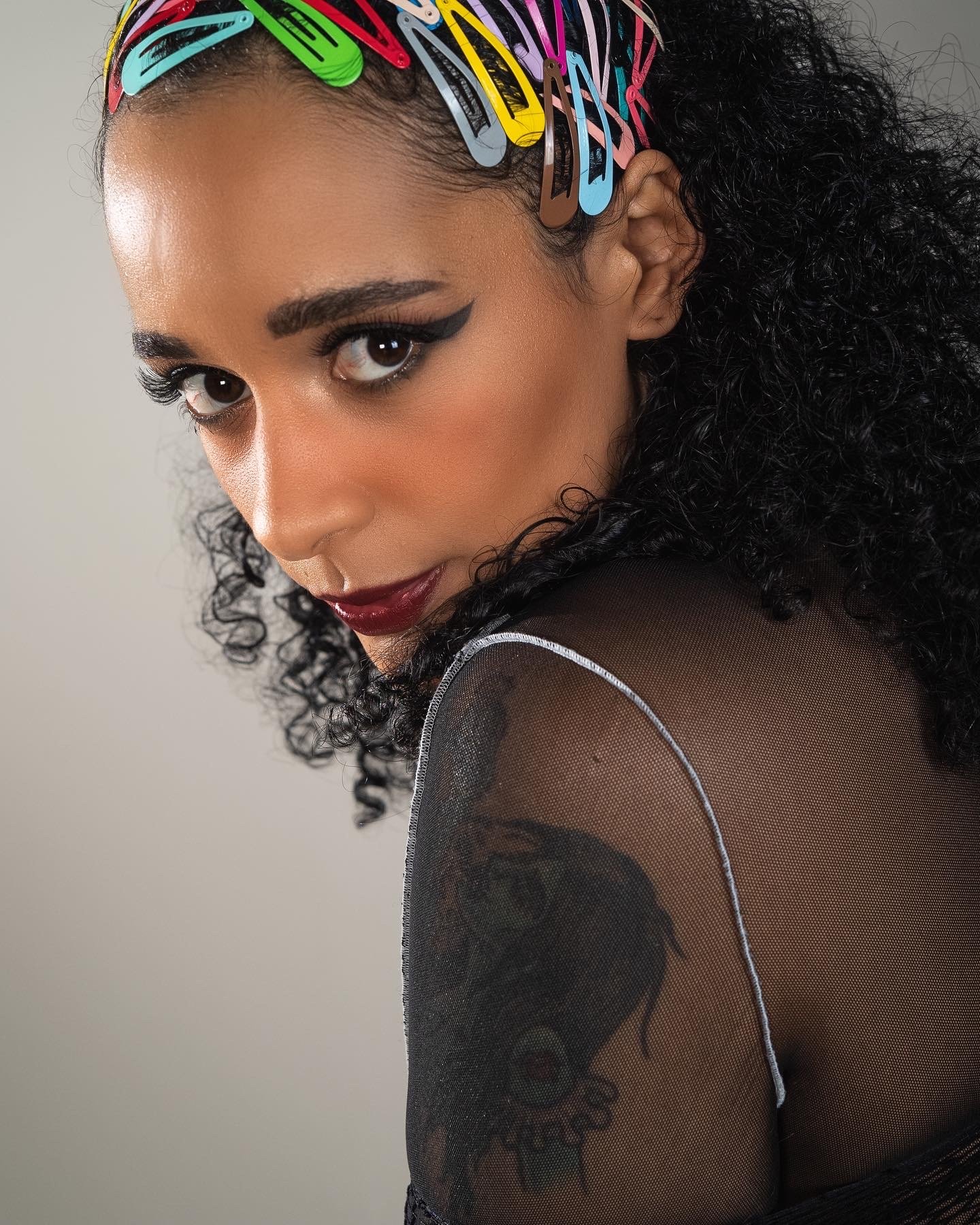 Close-up of a woman with curly black hair adorned with colorful hair clips, winged eyeliner, bold lipstick, and a sheer black top with visible tattoo on her shoulder.