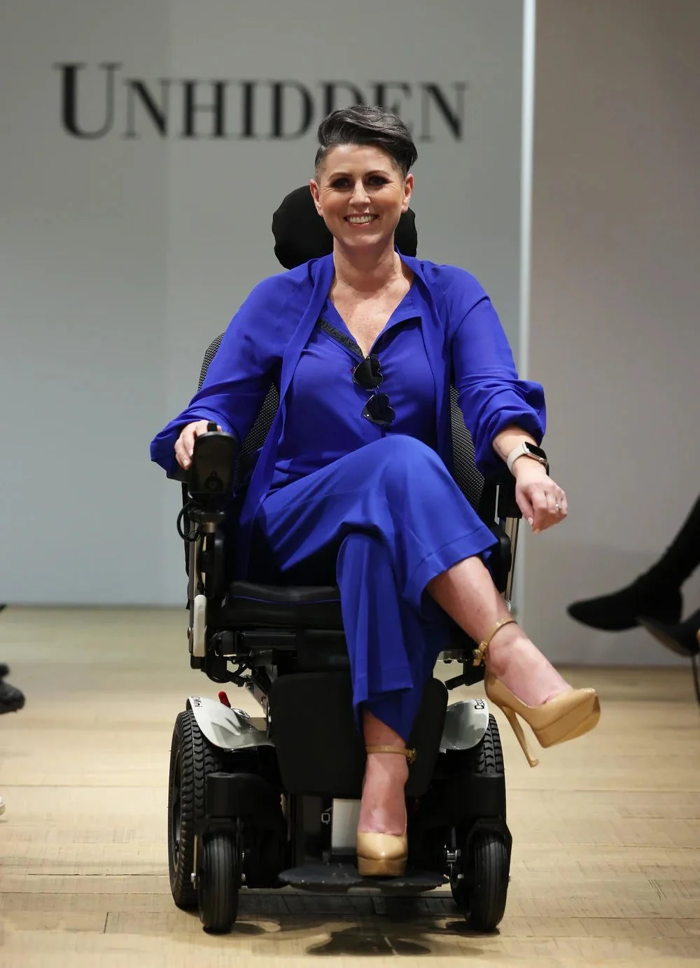 Woman in bright blue outfit sitting in a motorized wheelchair, smiling, with the word 'UNHIDDEN' in the background.