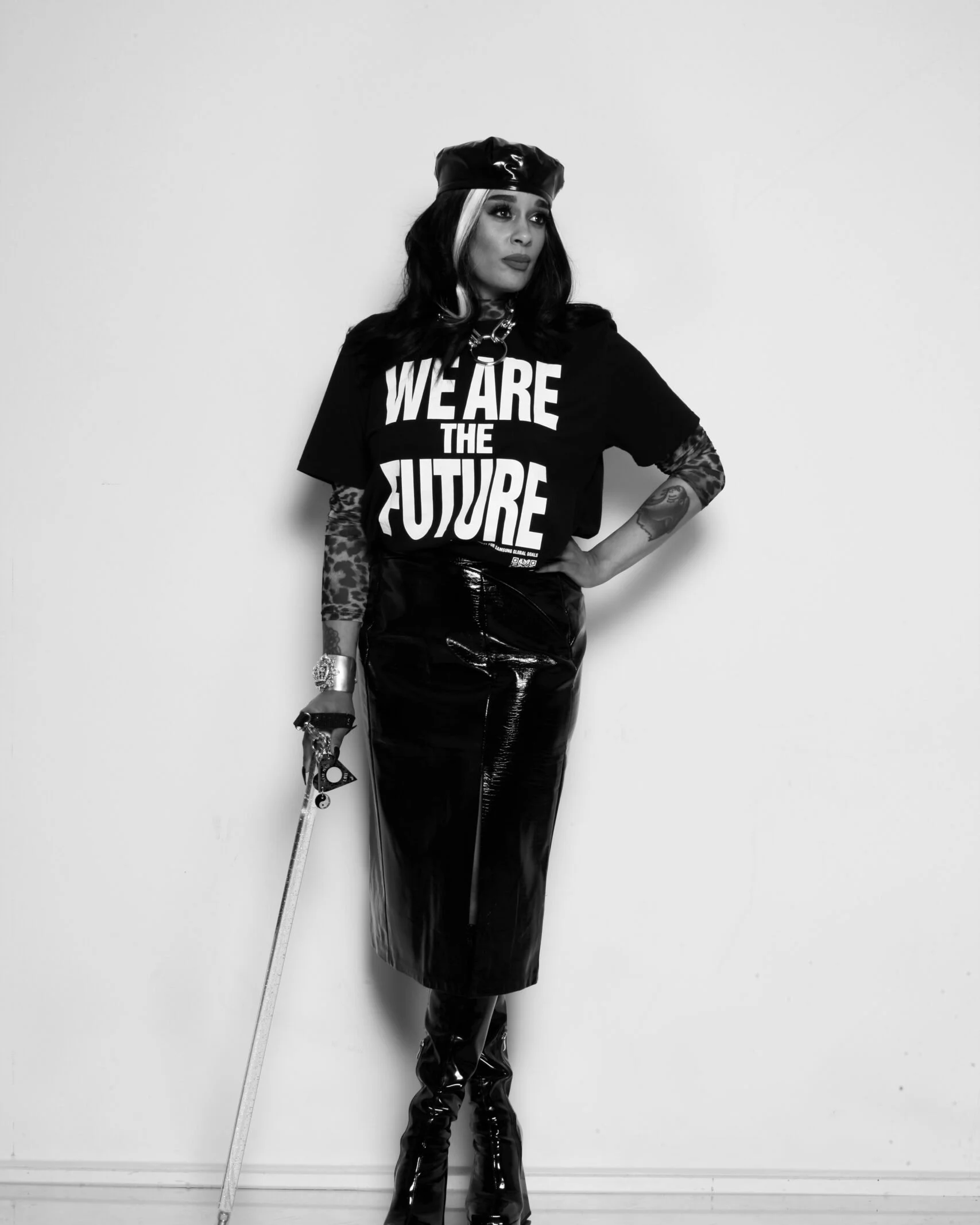 A woman standing against a plain wall, dressed in a bold fashion statement, wearing a black beanie, a t-shirt that reads 'We Are The Future,' a chain necklace, and shiny black pants and boots. She holds a cane and has tattoos on her arm.