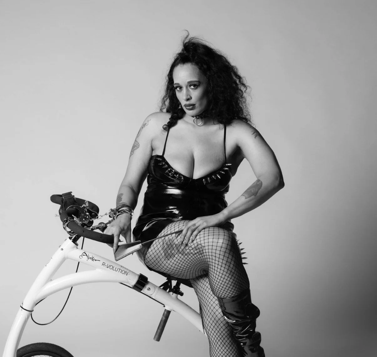A woman with curly hair and tattoos sitting on an electric bike, wearing a black shiny dress, fishnet stockings, and thigh-high boots, in a black and white studio portrait.