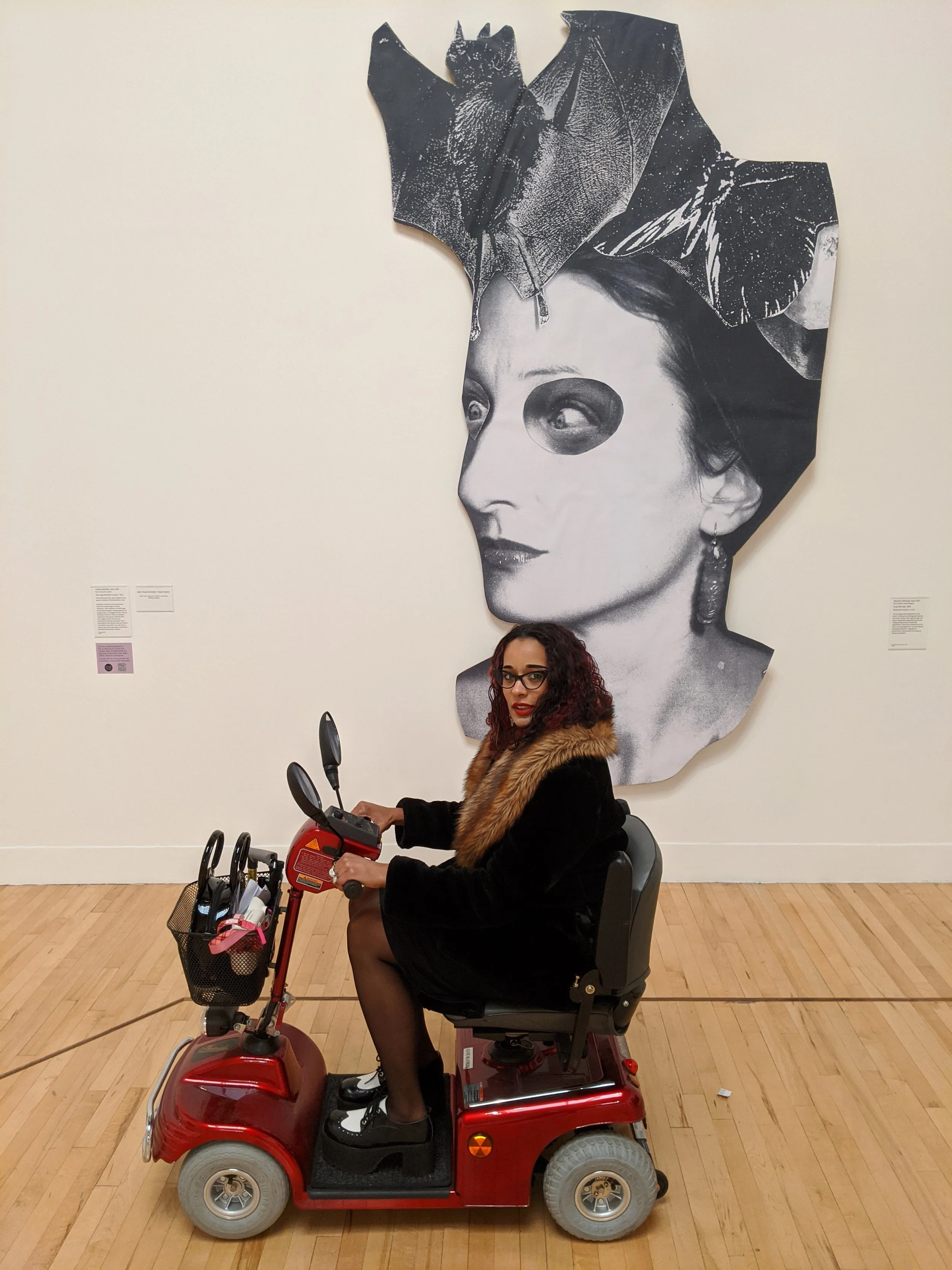 A woman sitting on a mobility scooter in front of a large black-and-white portrait of a woman with a leaf on her head in an art gallery.