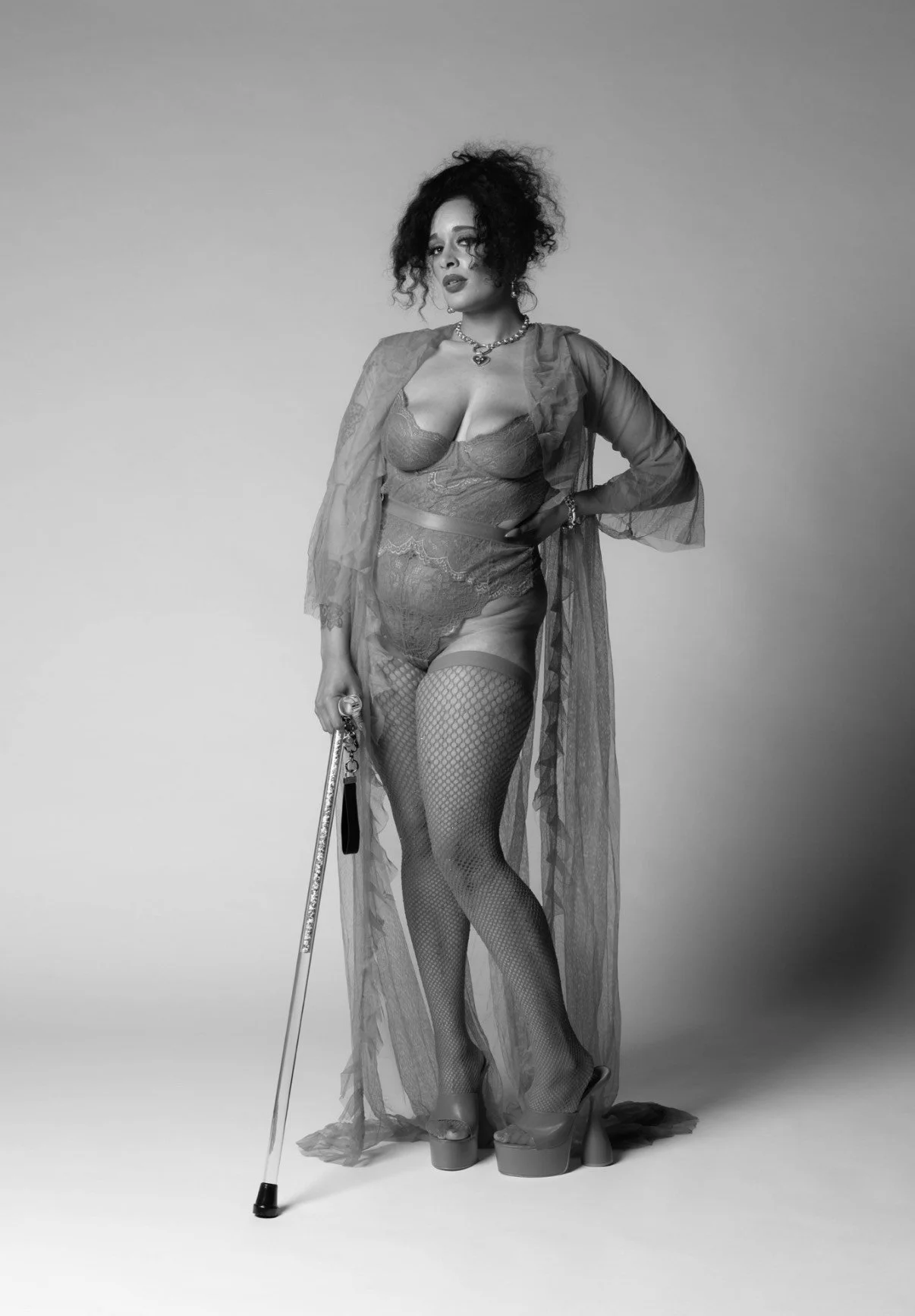 A woman with curly hair, wearing lace lingerie, stockings, and platform shoes, standing with a cane, in a studio with a plain background, in black and white.