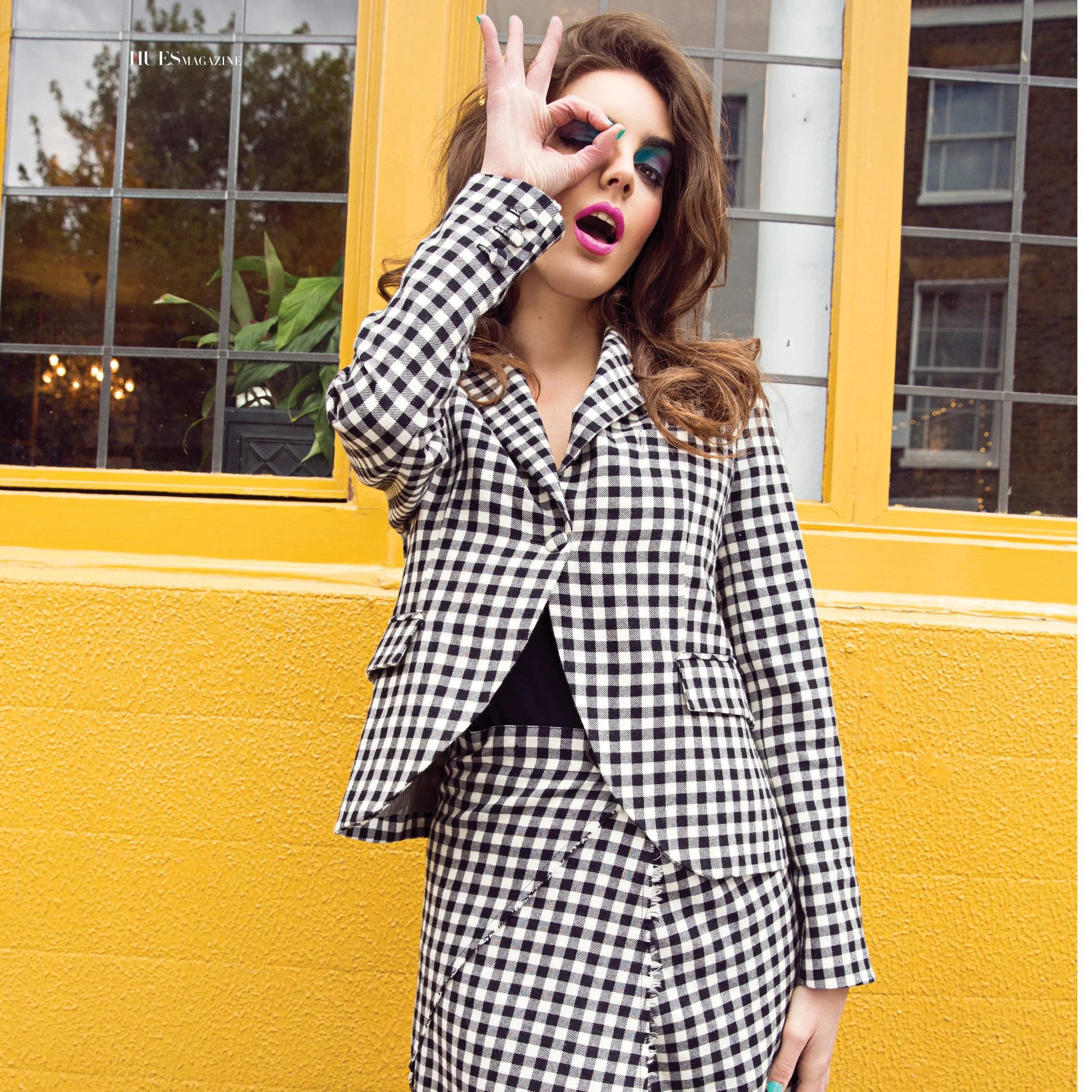 A woman with long wavy brown hair wearing a black and white checkered blazer standing in front of a bright yellow building with a grid window. She has colorful makeup, including teal eyeshadow and pink lipstick, and is making an 'okay' gesture with her right hand around her eye.