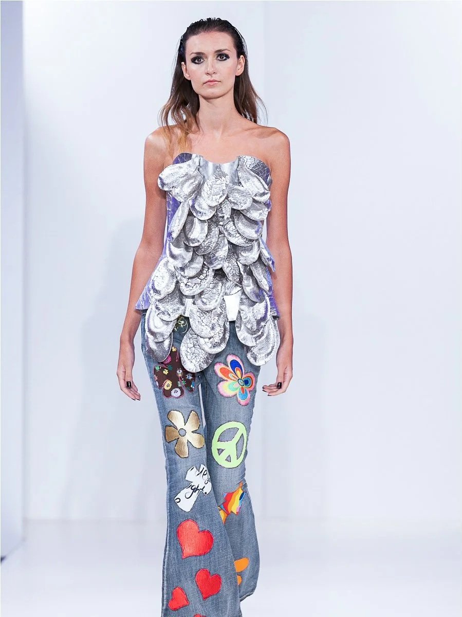 A woman standing against a plain white background, wearing a sleeveless dress with large silver ruffle embellishments on the front and pants decorated with colorful patches including hearts, a peace sign, and flowers.