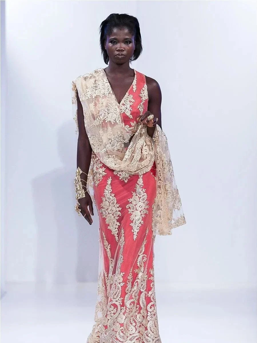 A woman with dark skin and short black hair wearing a pink and gold embroidered dress with a matching sheer scarf draped over her shoulder, standing against a plain white background.