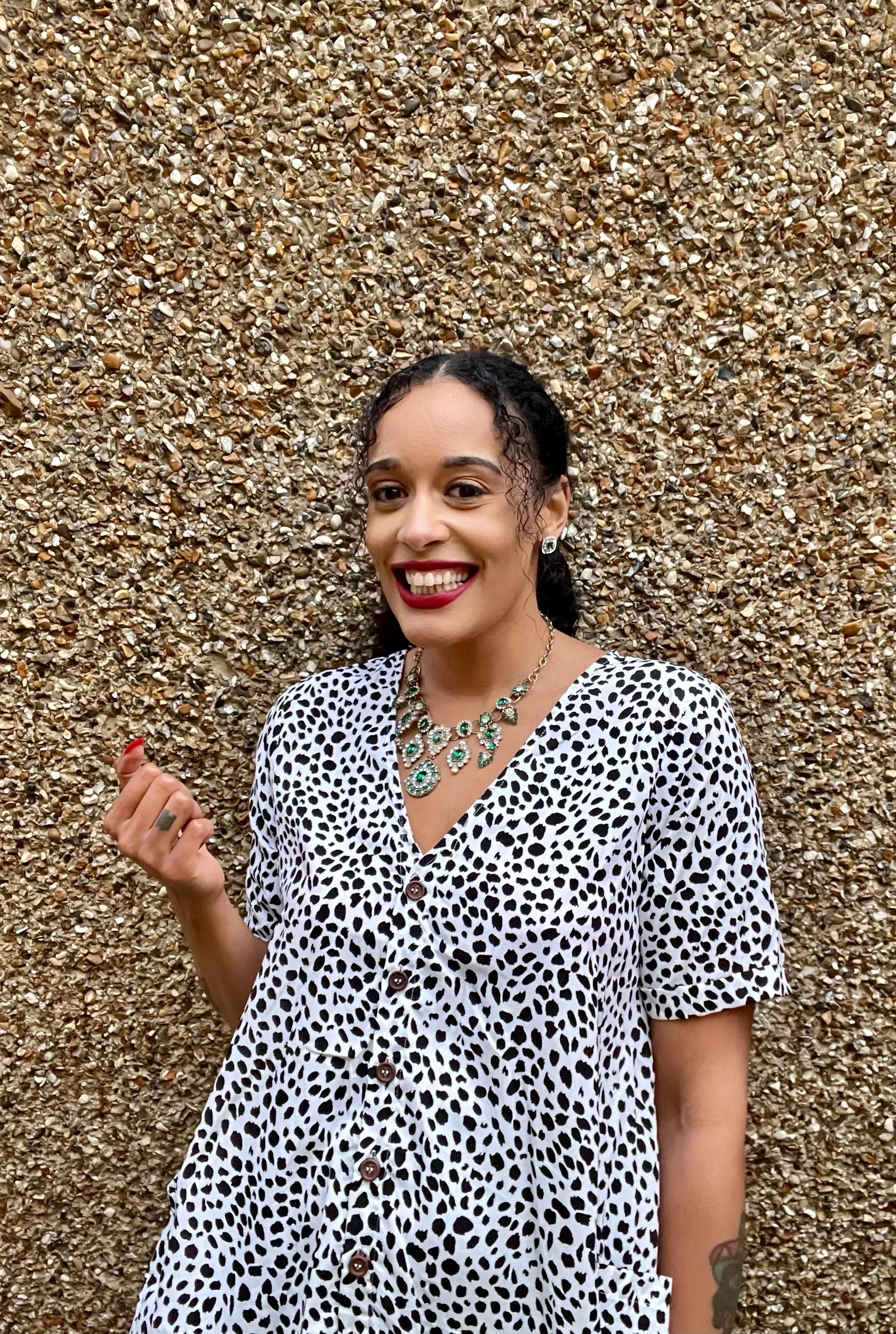 Smiling woman with curly black hair, wearing a leopard print dress and green gemstone jewelry, standing in front of a textured layer of small stones.