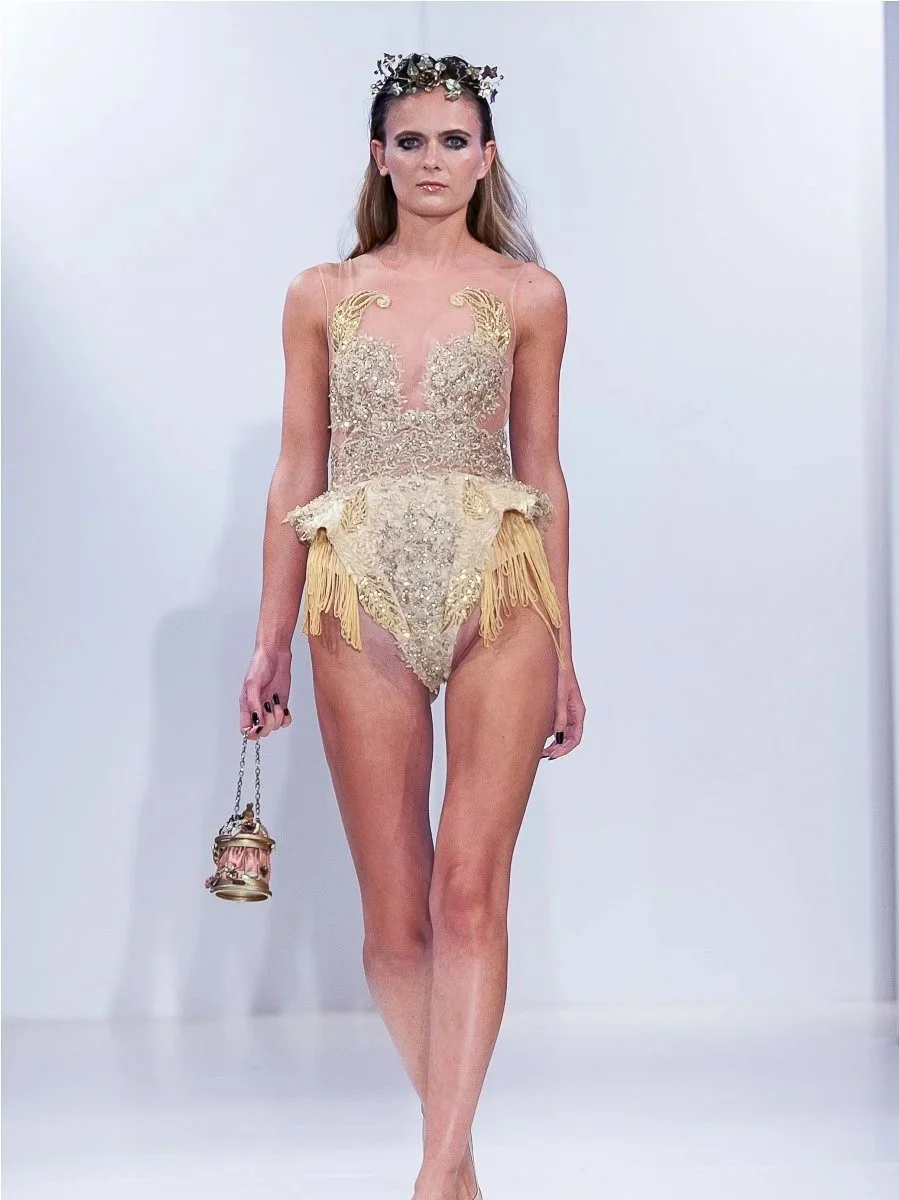 A model walking down the runway in a gold and beige ornate dress with fringe details. She has a floral headpiece and is carrying a small decorative purse in her right hand.
