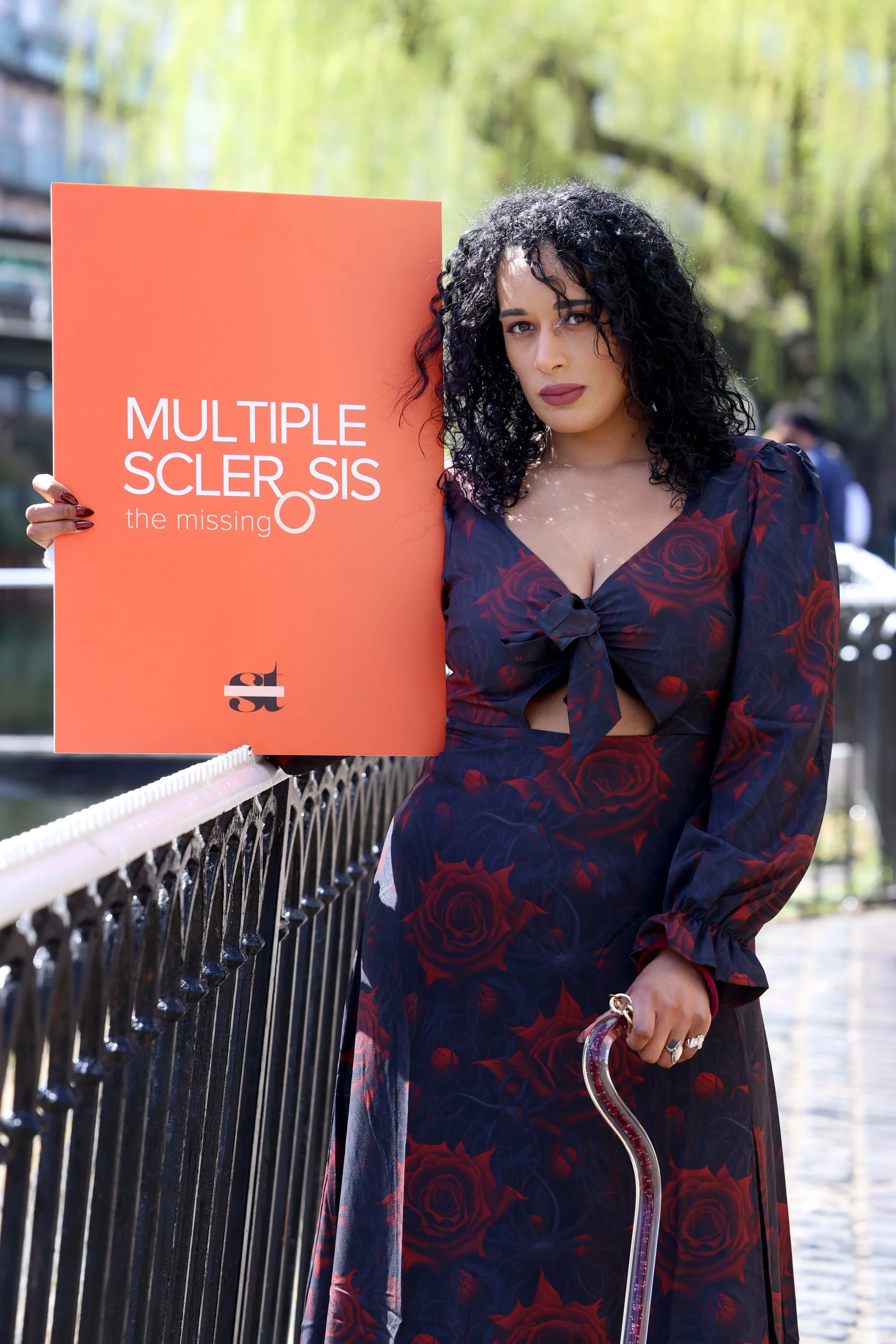 A woman with dark curly hair wearing a dark floral dress holding an orange sign with the text "Multiple sclerosis the missing O" and a black and white logo underneath, standing by a black metal fence outdoors with trees in the background.