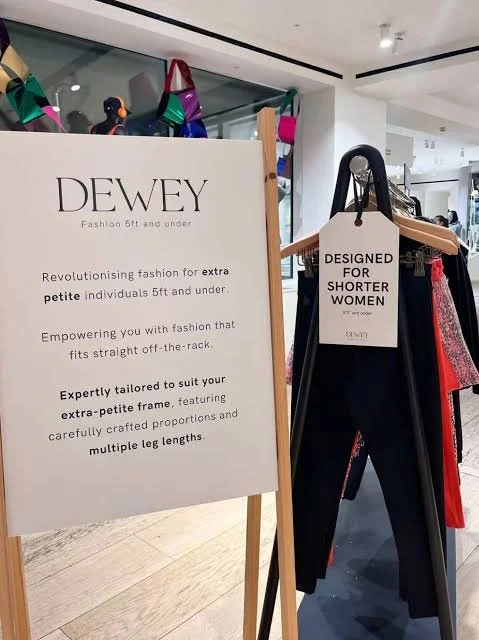 Clothing display in a store with an informational sign and a rack of clothes, including pants and dresses, designed for shorter women under the Dewey brand.