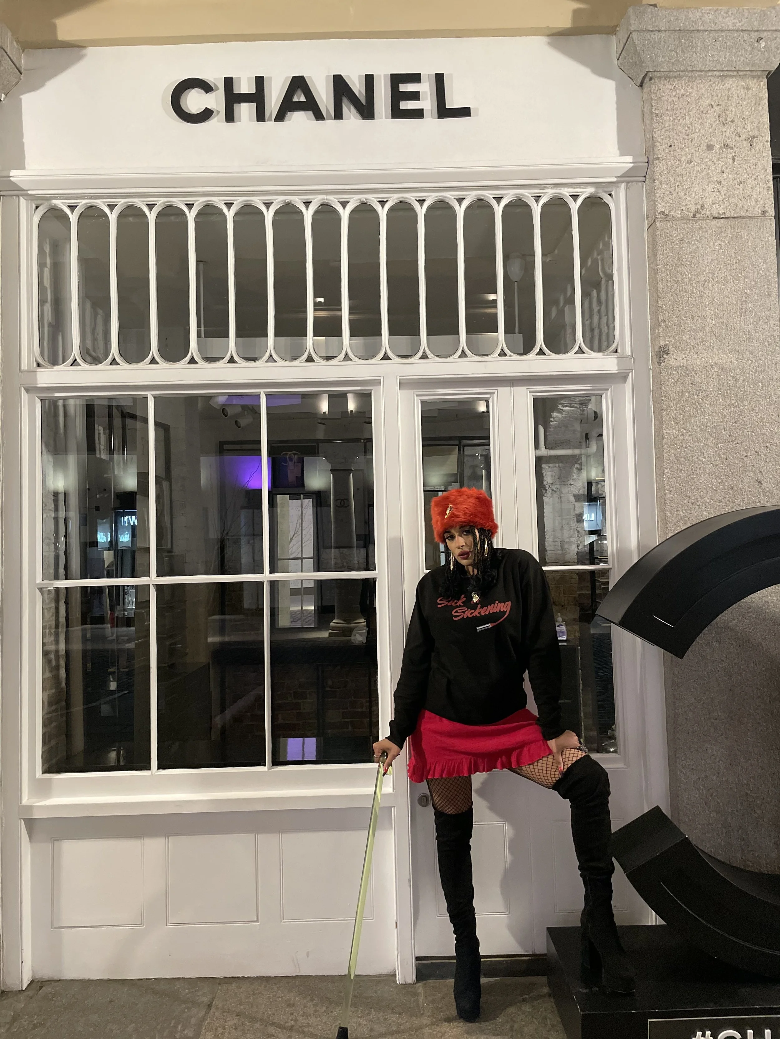 Person wearing a red fur hat, black hoodie with red text, red skirt, fishnet stockings, and thigh-high boots standing in front of a store called Chanel.