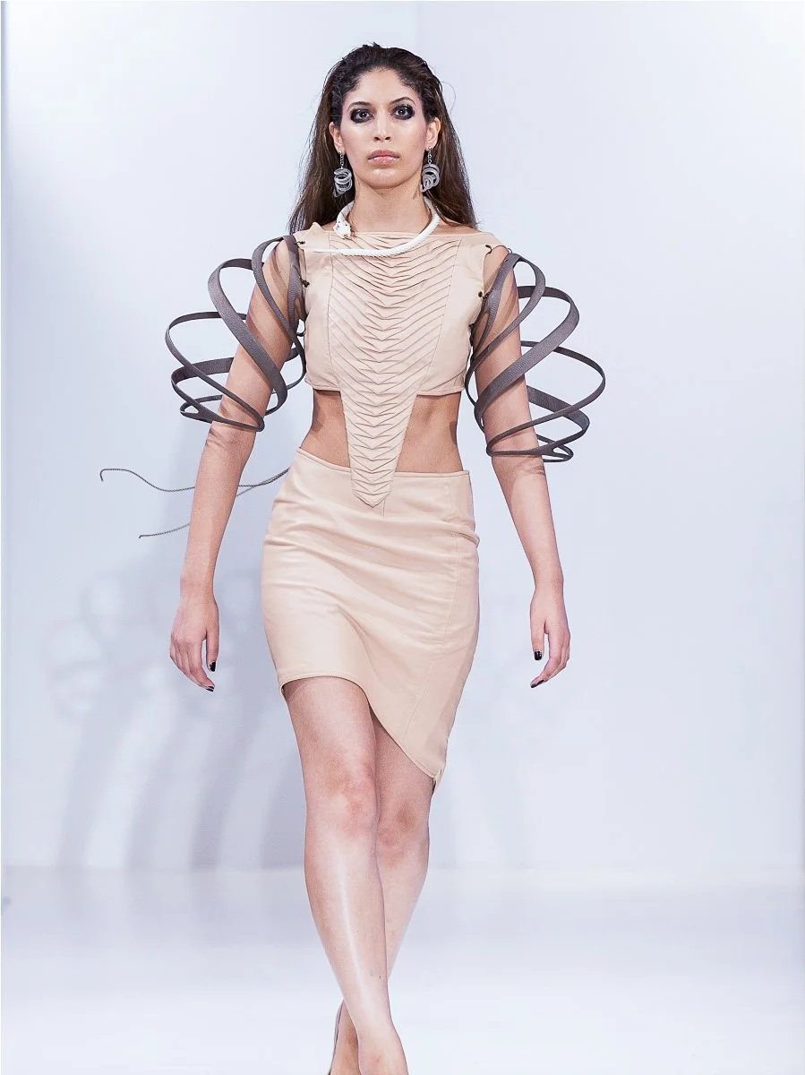 A female model walking on a runway in a beige, modern, and artistic dress with unique, curly sleeve accents.