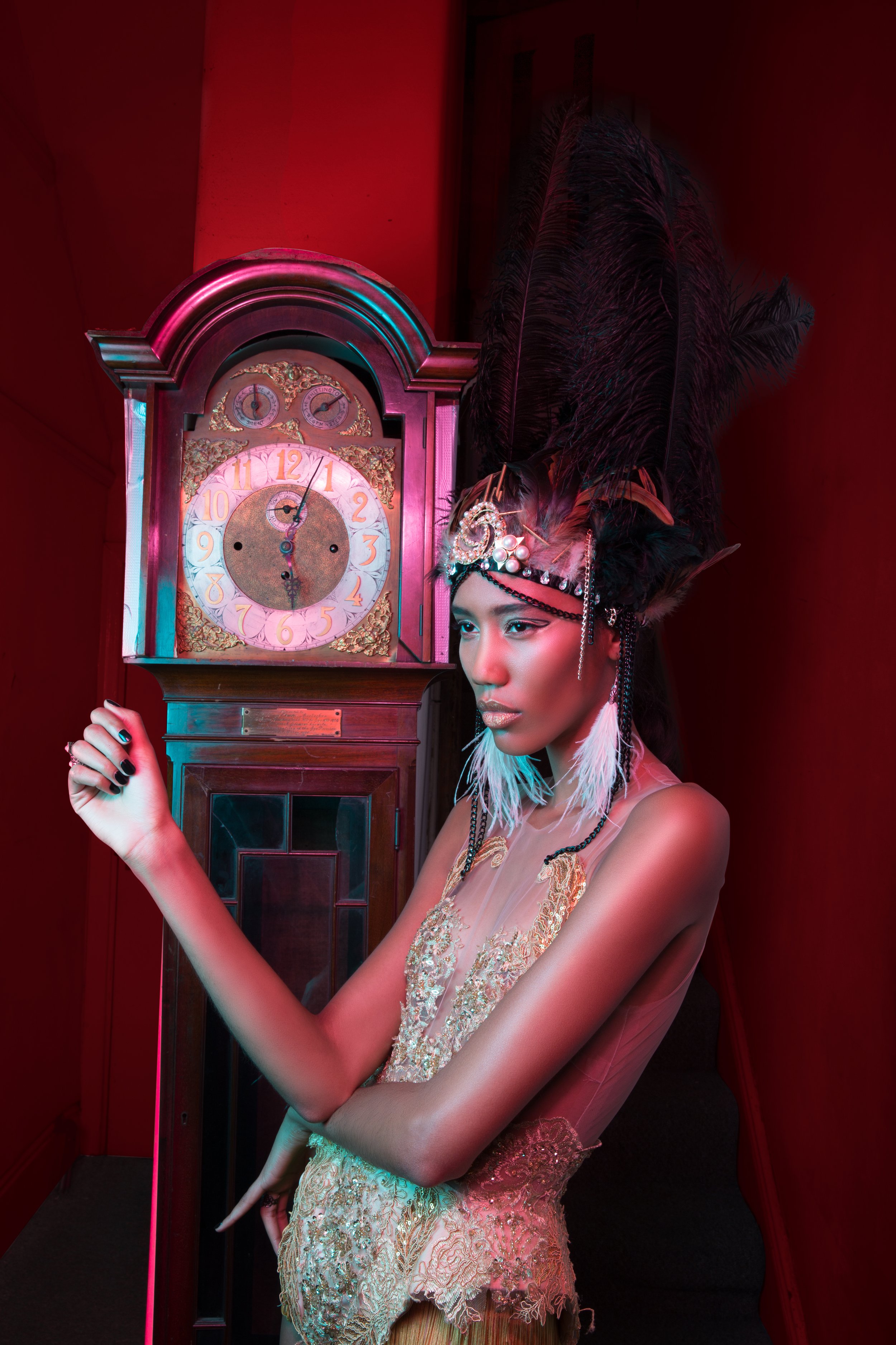 A woman with dark skin pose in front of a vintage clock and red wall, wearing an ornate headdress with large black feathers and decorations, and a sheer gold embroidered dress.