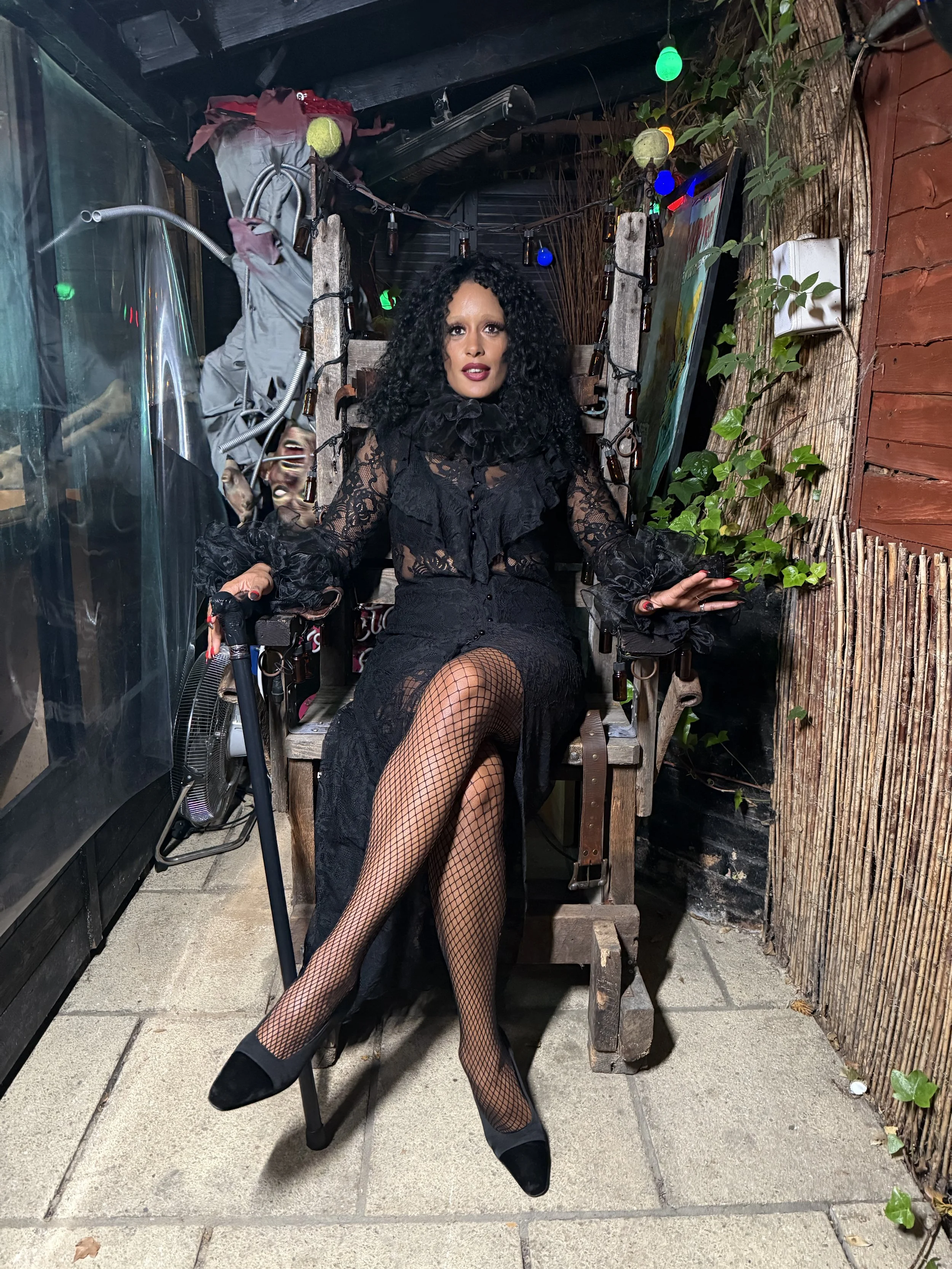 A woman with curly black hair, wearing a black lace dress with puffed sleeves and fishnet stockings, sits on a wooden chair in a decorated outdoor patio area at night. She has makeup on and is posing with one leg crossed over the other, hands resting on the arms of the chair. The background has colorful string lights, a wooden fence with green ivy, and some paintings.