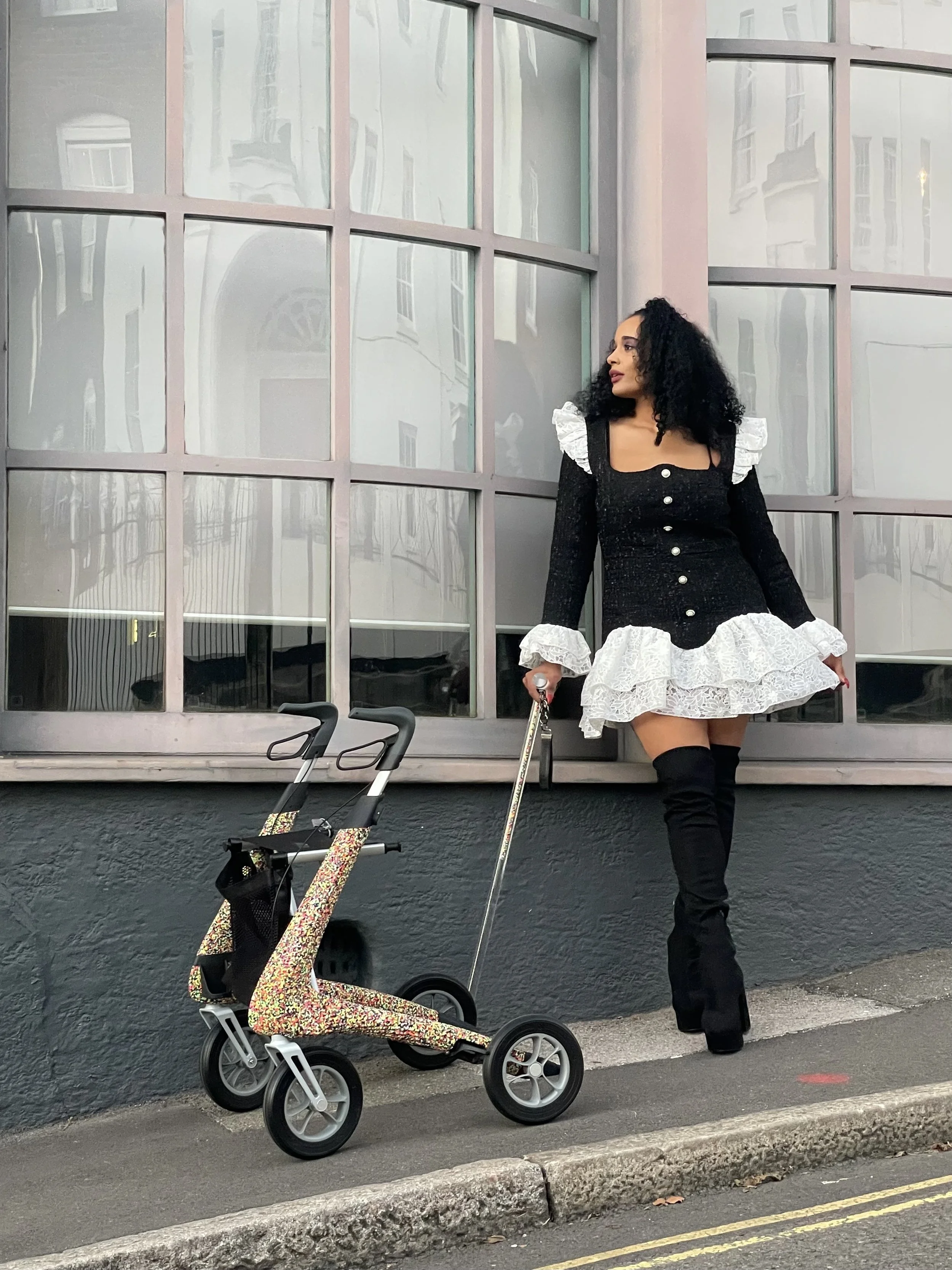 A woman with curly black hair wearing a black and white dress and black thigh-high boots standing on a sidewalk next to a black wall. She is holding a walking cane. An empty stroller with a floral pattern is in front of her.