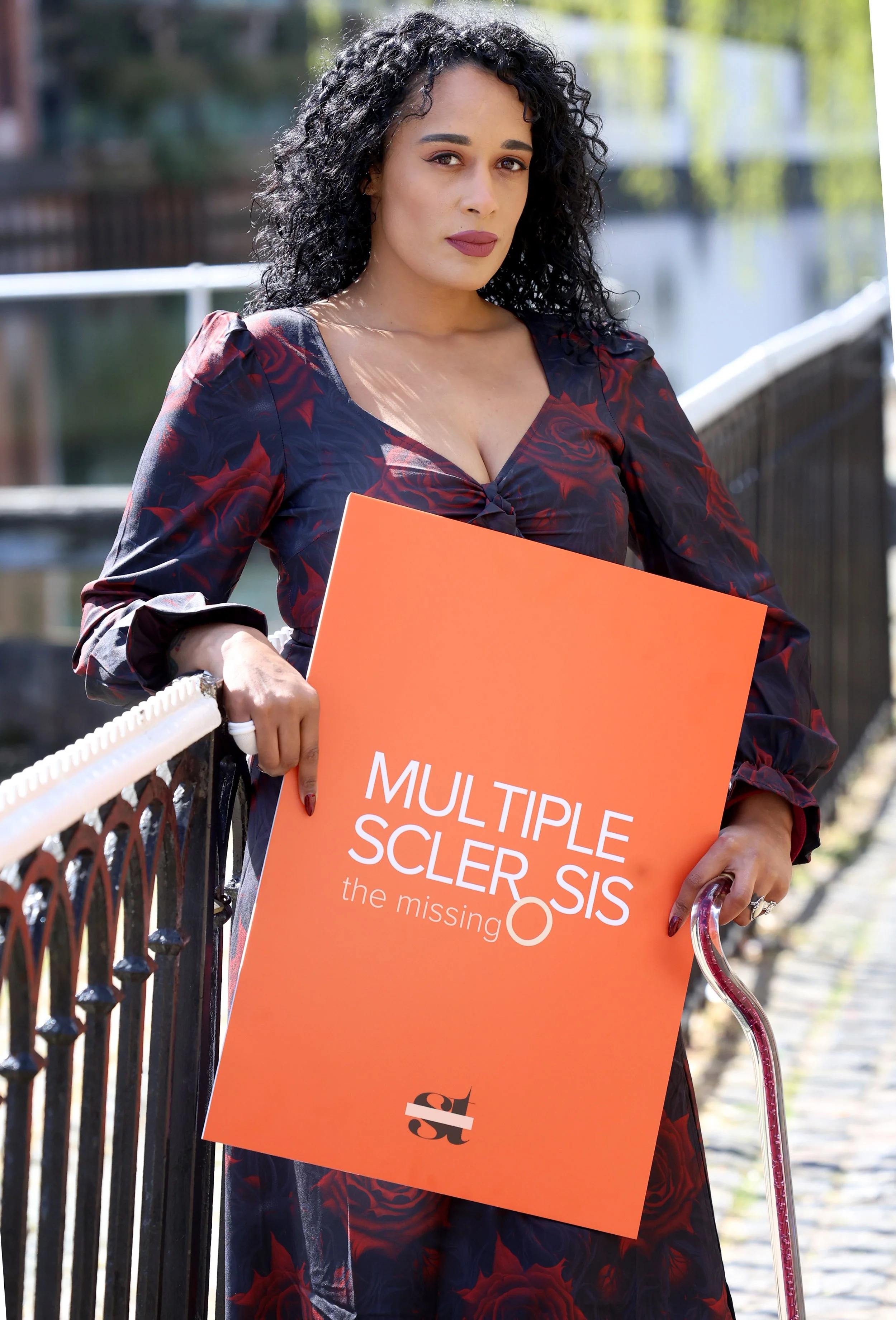 A woman with curly black hair holding a protest sign that reads 'Multiple Sclerosis Sis: the missing part'. She is standing outdoors next to a black iron fence, with a canal and trees in the background.