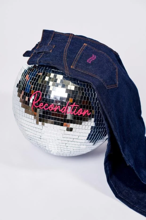 Pair of dark blue jeans draped over a disco ball with the word 'Recondition' written in pink on the ball.