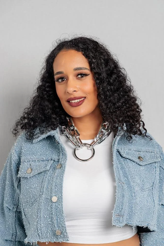 A woman with curly black hair wearing a chunky silver chain necklace, a white top, and a light blue denim jacket with metal buttons, smiling at the camera against a plain gray background.