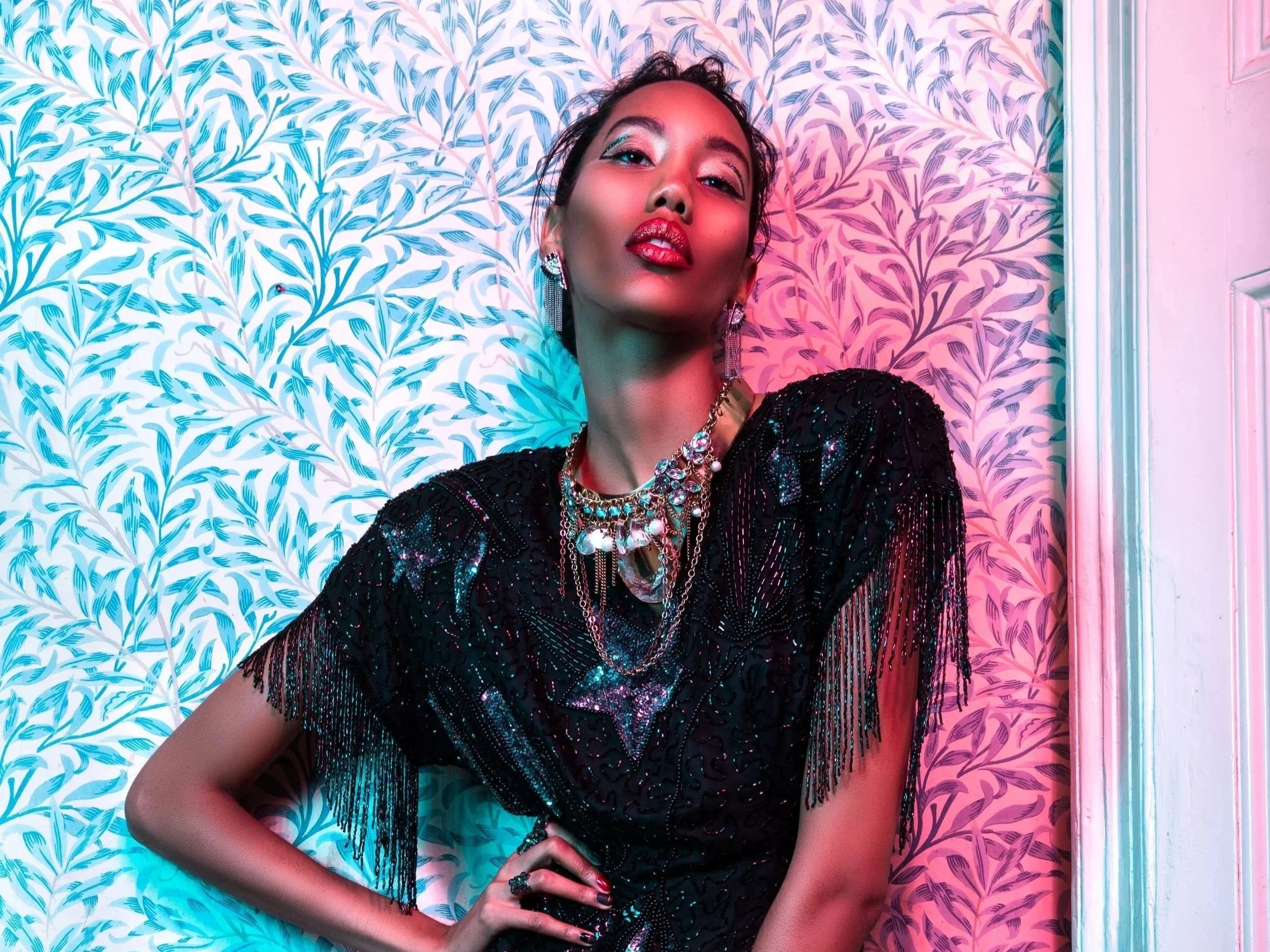 A woman with dark, wavy hair and bold makeup, including red lipstick and eye makeup, lying against a colorful, patterned wall. She is wearing a black, shiny, fringed dress and layered jewelry with pearls and crystals.