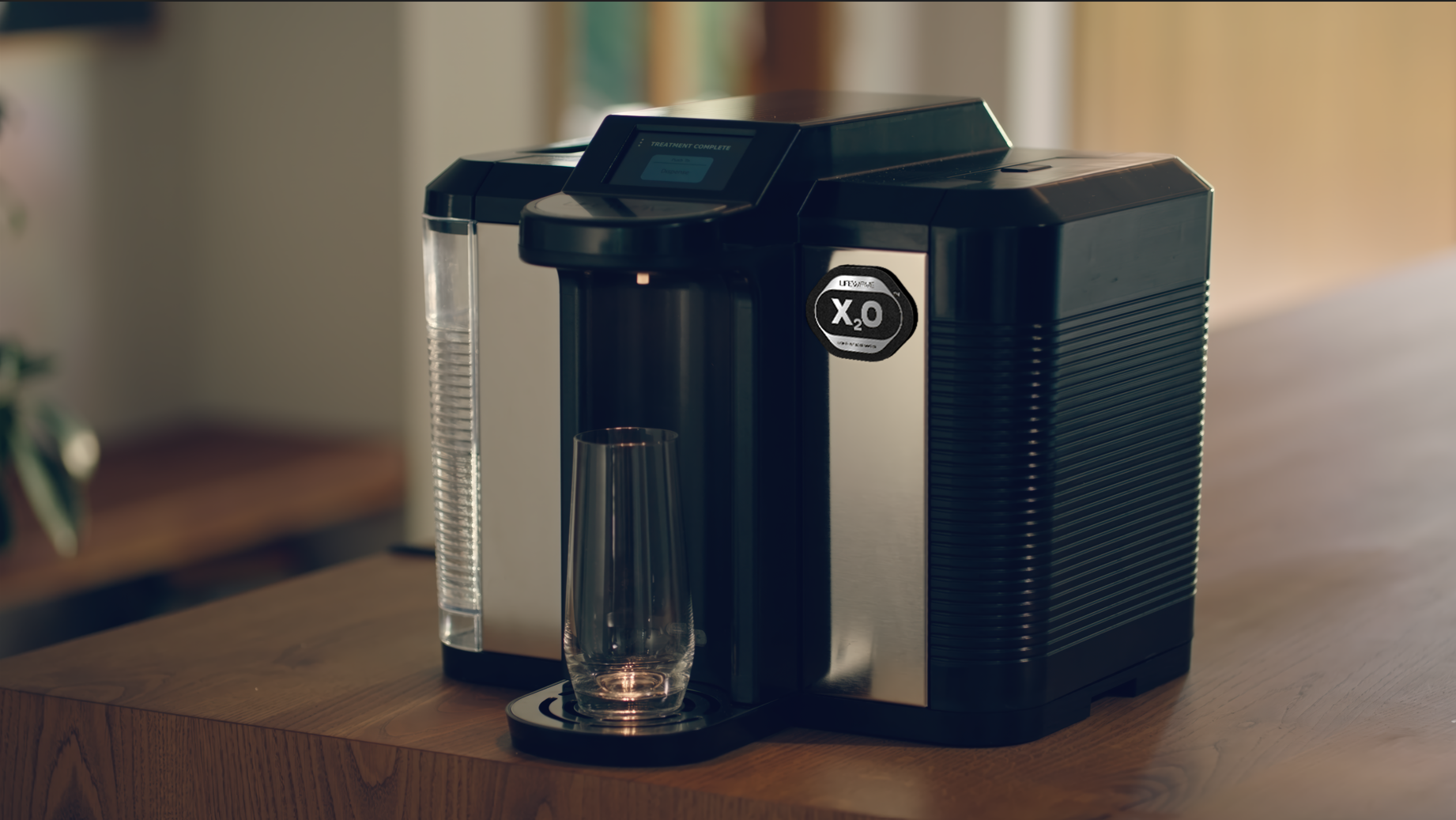 X2O system with a transparent water reservoir, a glass cup placed on a drip tray on a wooden table.