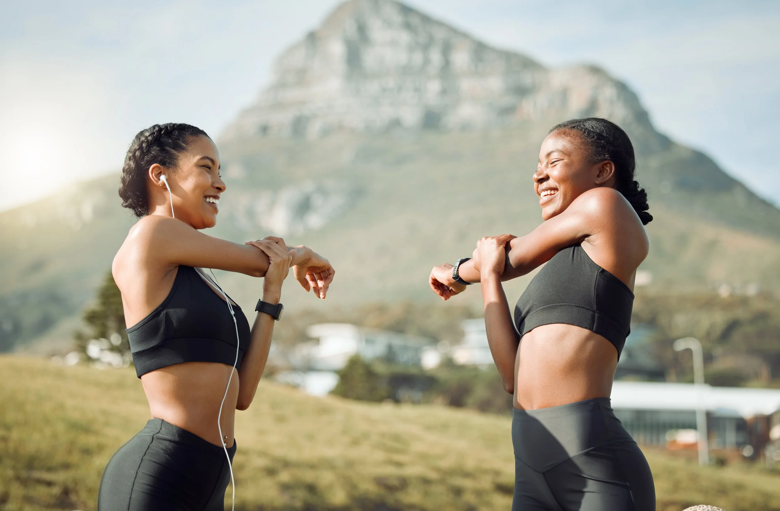 Two women in athletic wear stretching outdoors with a mountain in the background, smiling and facing each other.