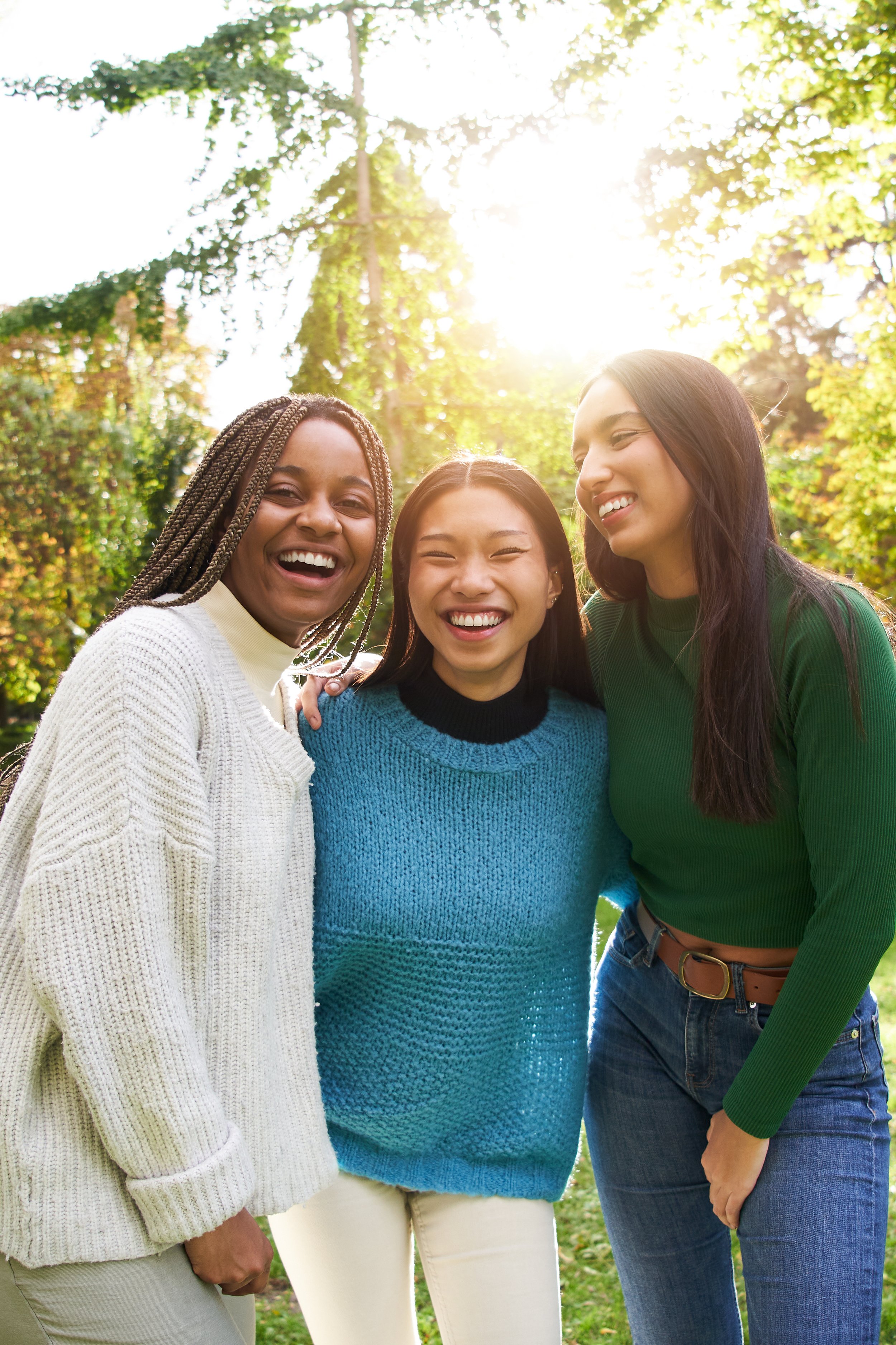 Three women standing outdoors in sunlight, smiling, with trees and greenery in the background.