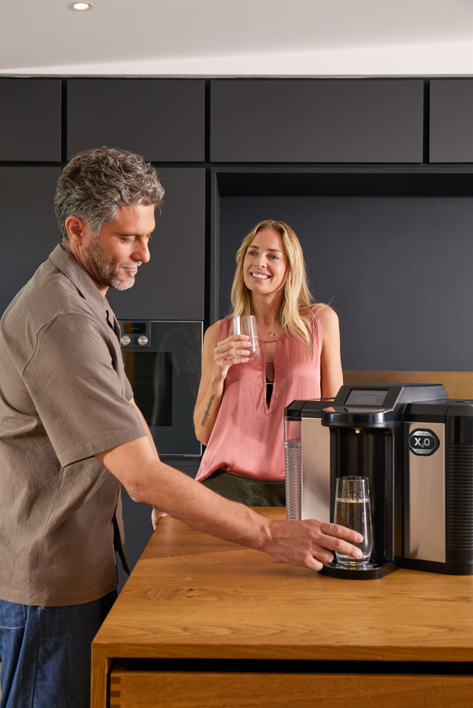 A man and a woman in a modern kitchen, with the man pouring water from an X2O water dispenser into a glass, while the woman stands nearby holding a glass and smiling.