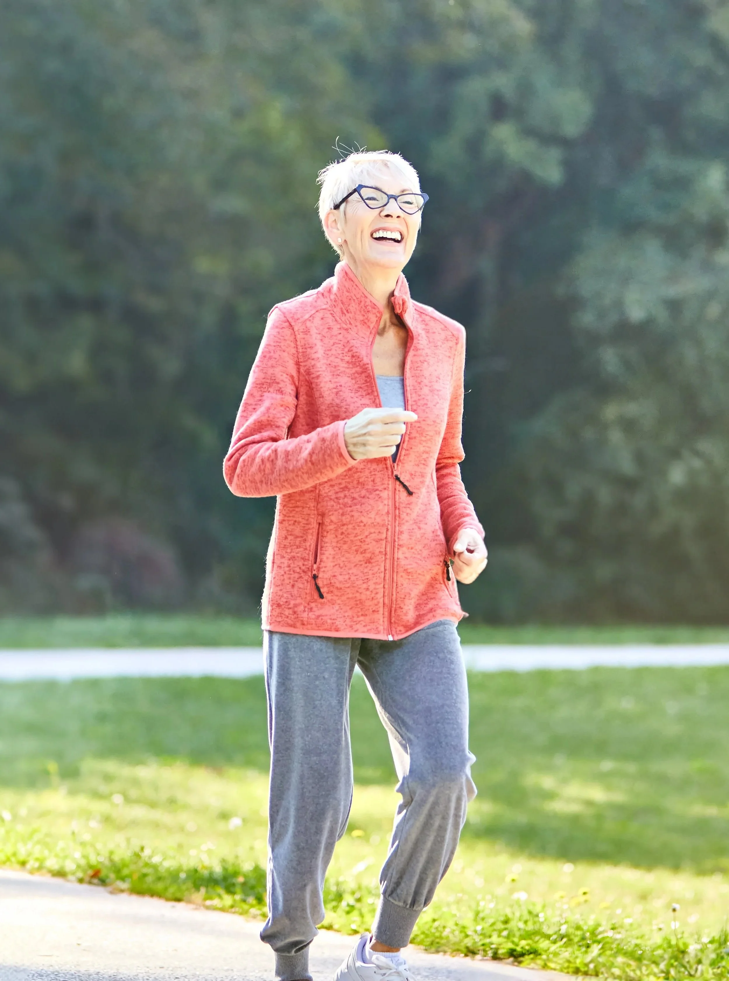 An elderly woman jogging outdoors in a park, smiling, wearing glasses, a coral-colored jacket, grey sweatpants, and white sneakers.